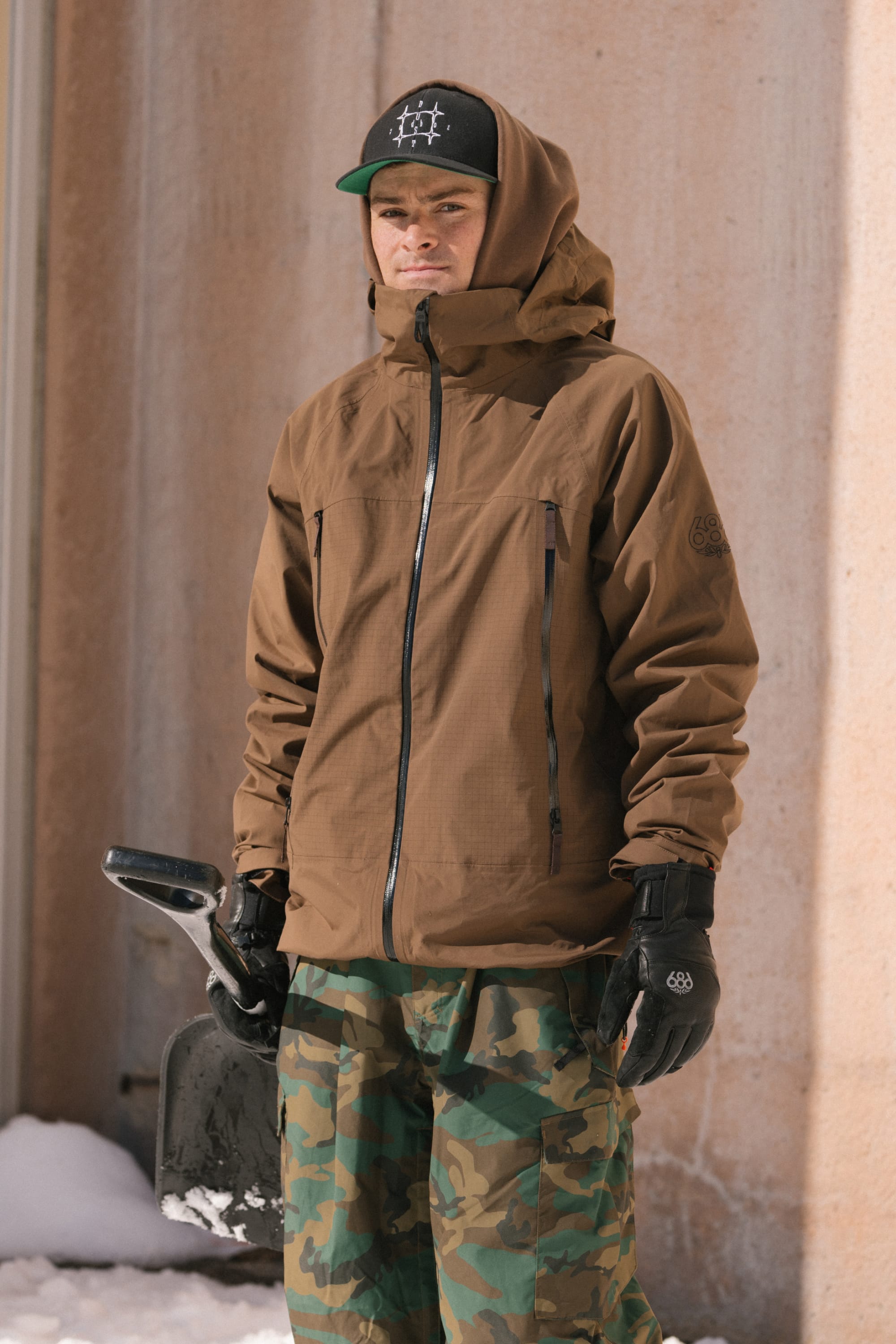 A person wearing the 686 Men’s SMARTY® 3-In-1 Rodeo™ 3L Jacket by 686, along with camouflage pants and black gloves, stands outdoors near a wall with snow at their feet, holding a snow shovel. They are also wearing a black cap.