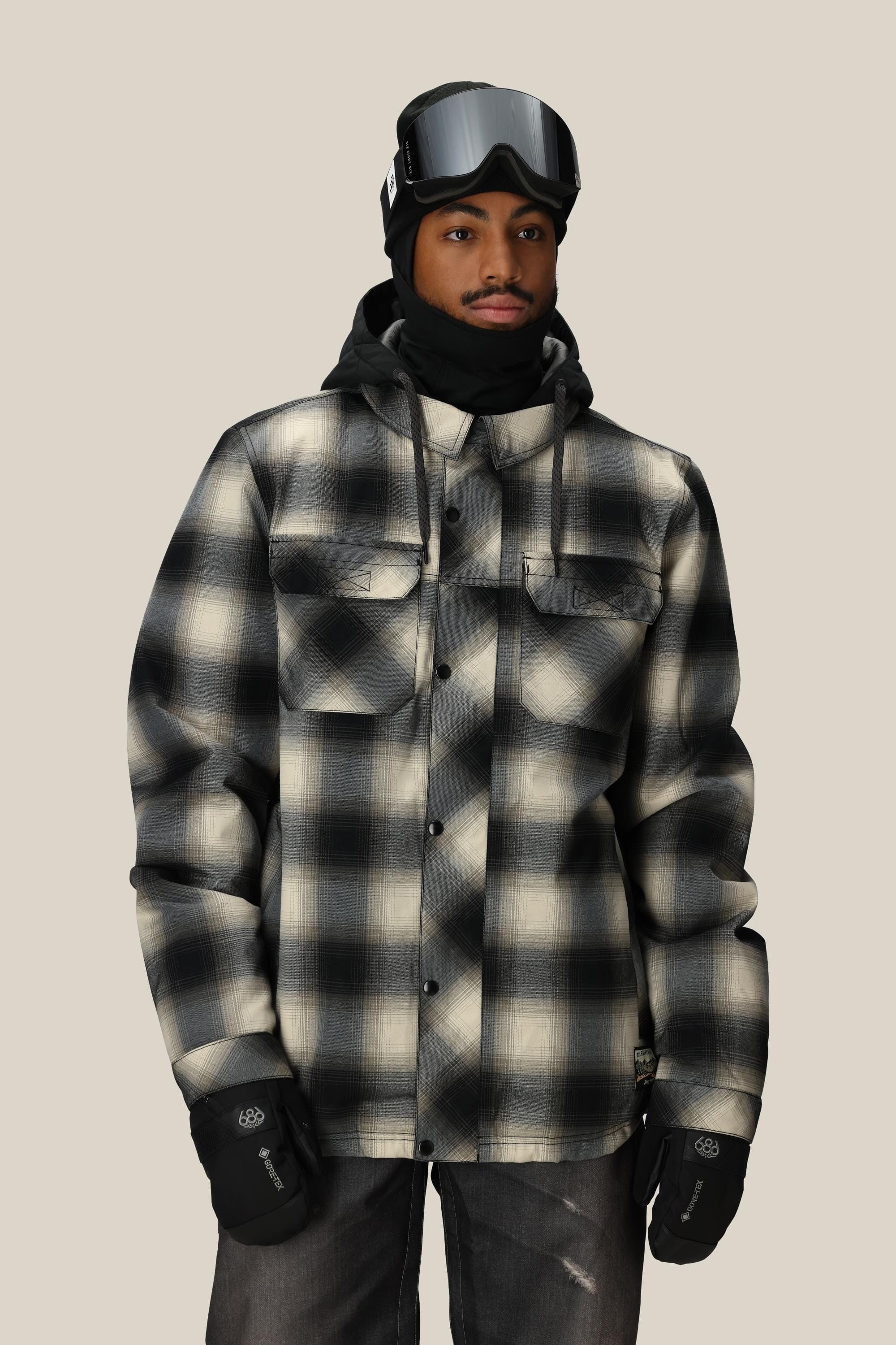 A person stands facing the camera against a plain beige background, wearing the 686 Men’s Woodland™ Insulated Jacket from 686 in a black-and-white plaid pattern, paired with black ski goggles, gloves, a black hood, and neck gaiter.