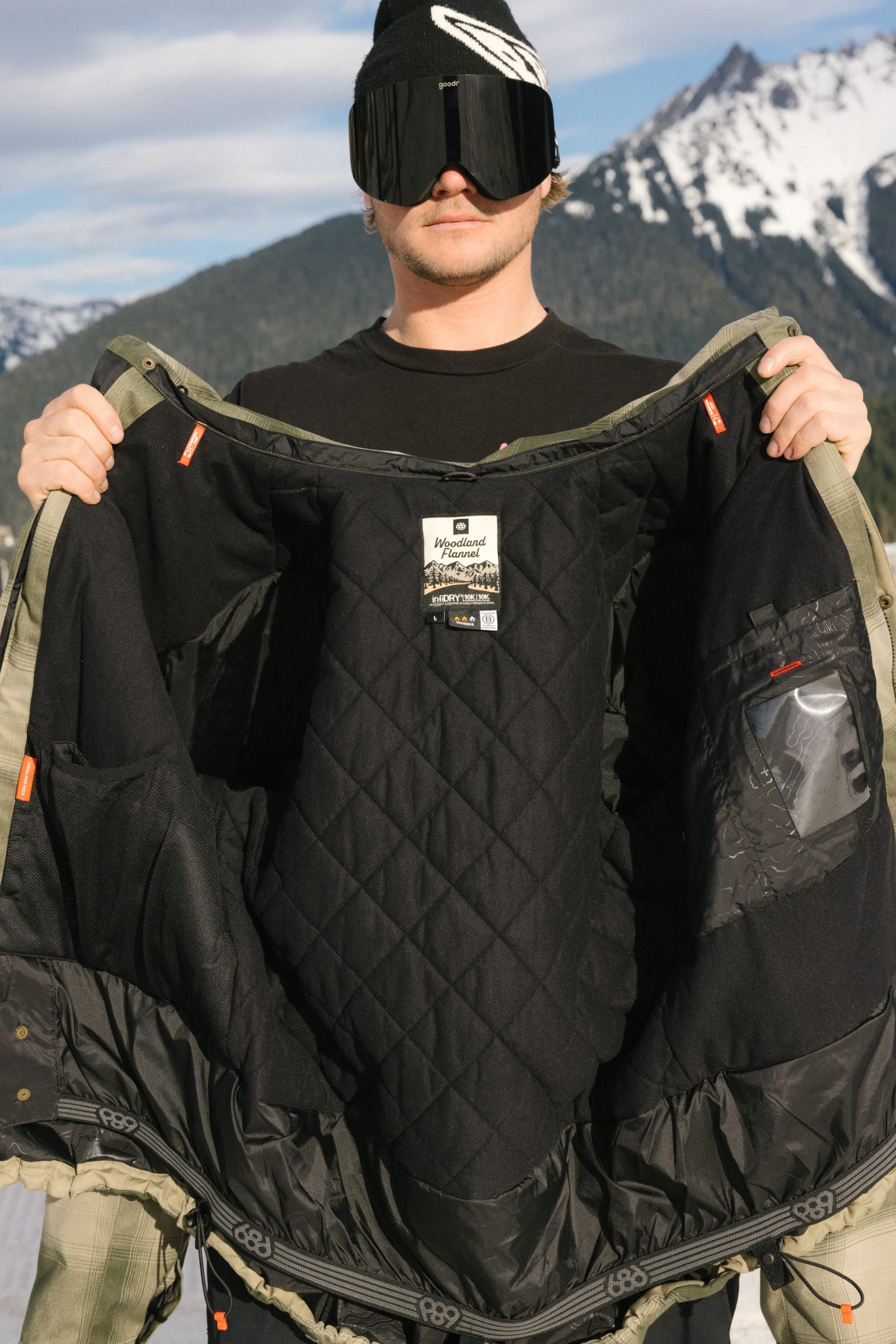 A person in ski gear holds open the 686 Men’s Woodland™ Insulated Jacket, showcasing its insulated interior lining against a backdrop of snow-covered mountains and blue sky.