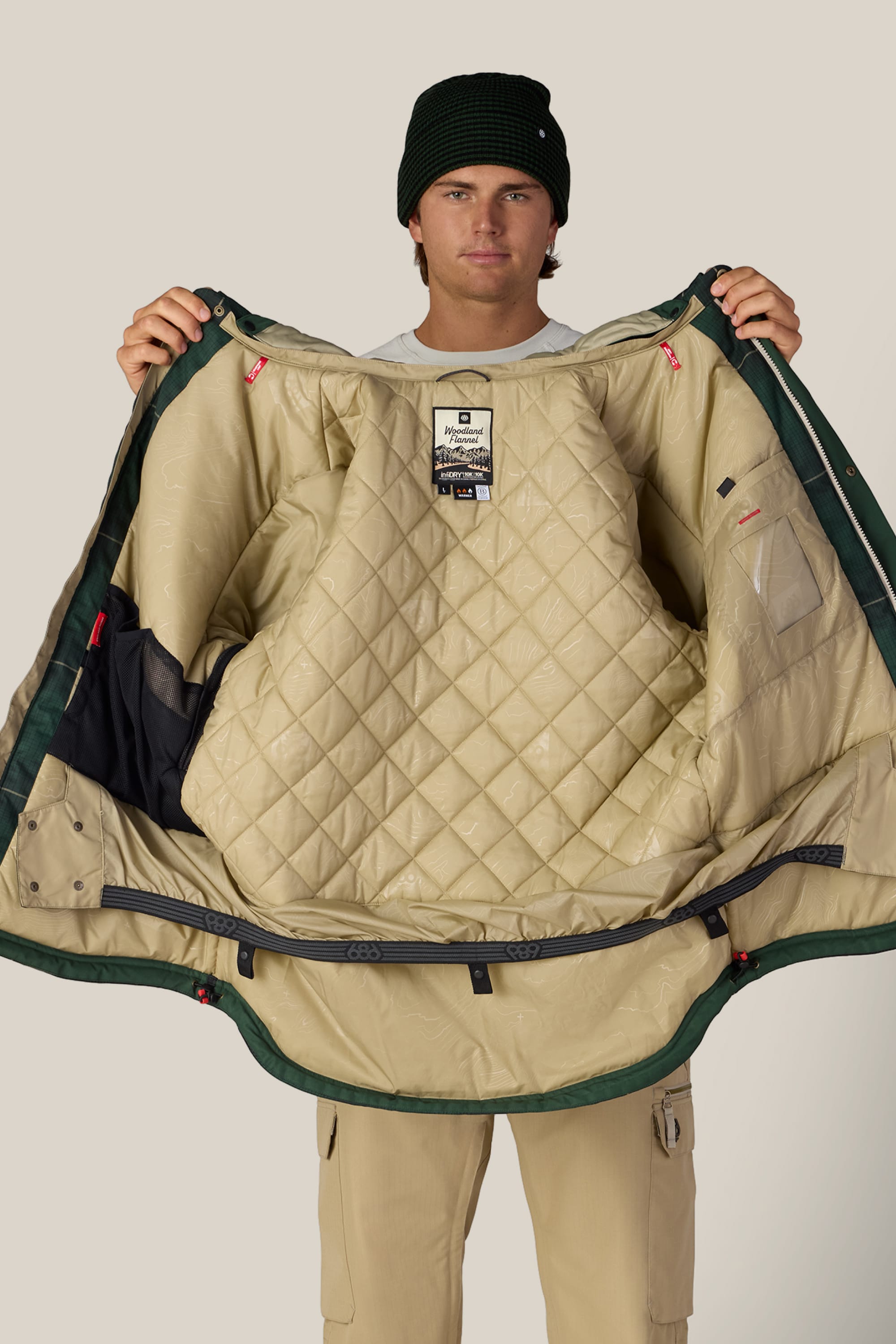 A man wearing a black beanie and beige outfit holds open the 686 Men’s Woodland™ Insulated Jacket, displaying its quilted tan exterior, lined interior, and multiple pockets against a neutral background.