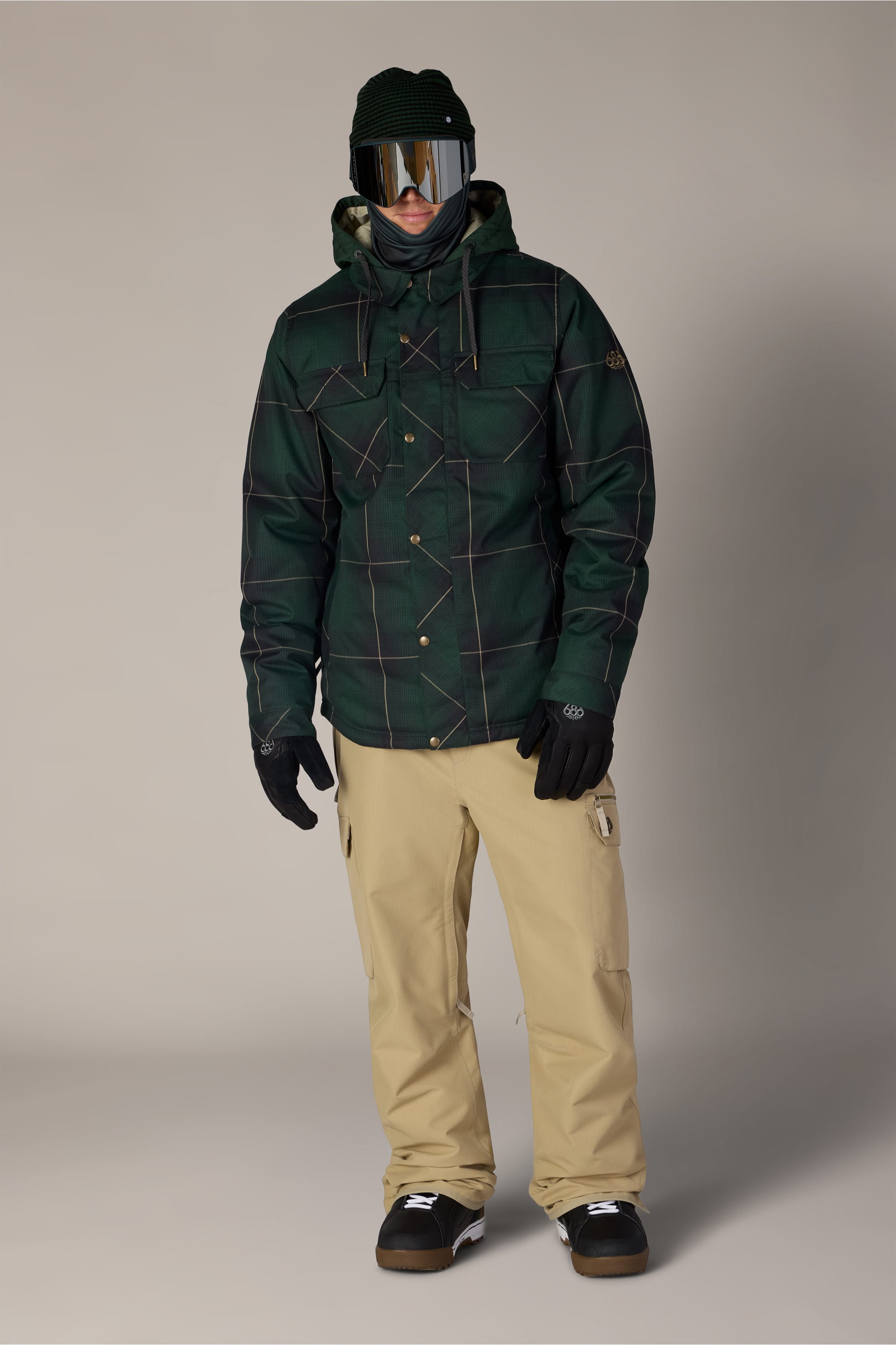 A person wearing the 686 Men’s Woodland™ Insulated Jacket by 686, along with beige snow pants, black gloves, a black beanie, and ski goggles, stands facing forward against a plain background.