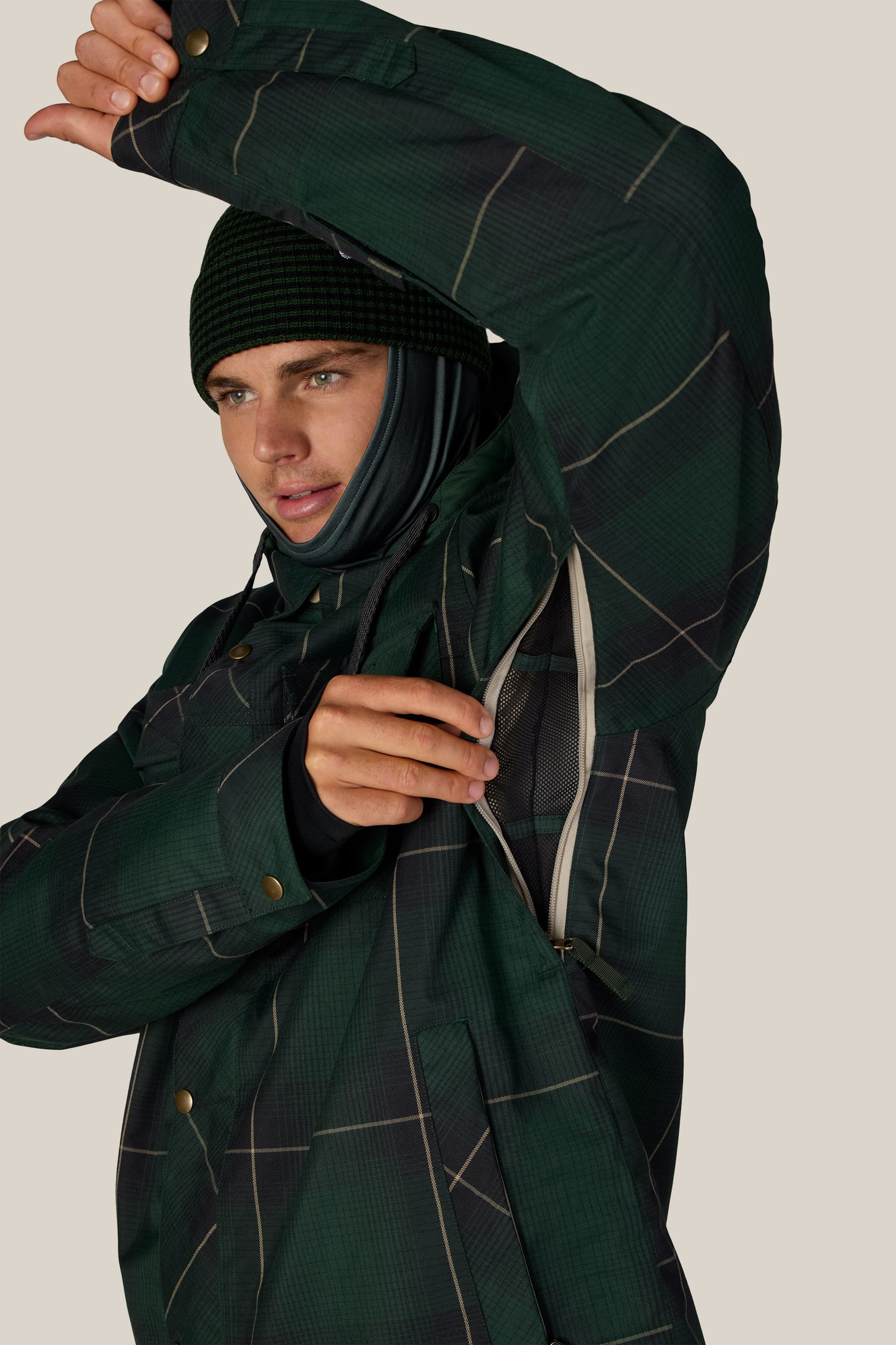 A person wearing the 686 Men’s Woodland™ Insulated Jacket in green plaid, paired with a black beanie, demonstrates the jacket’s underarm ventilation feature by unzipping it. The scene has a plain, light-colored background.