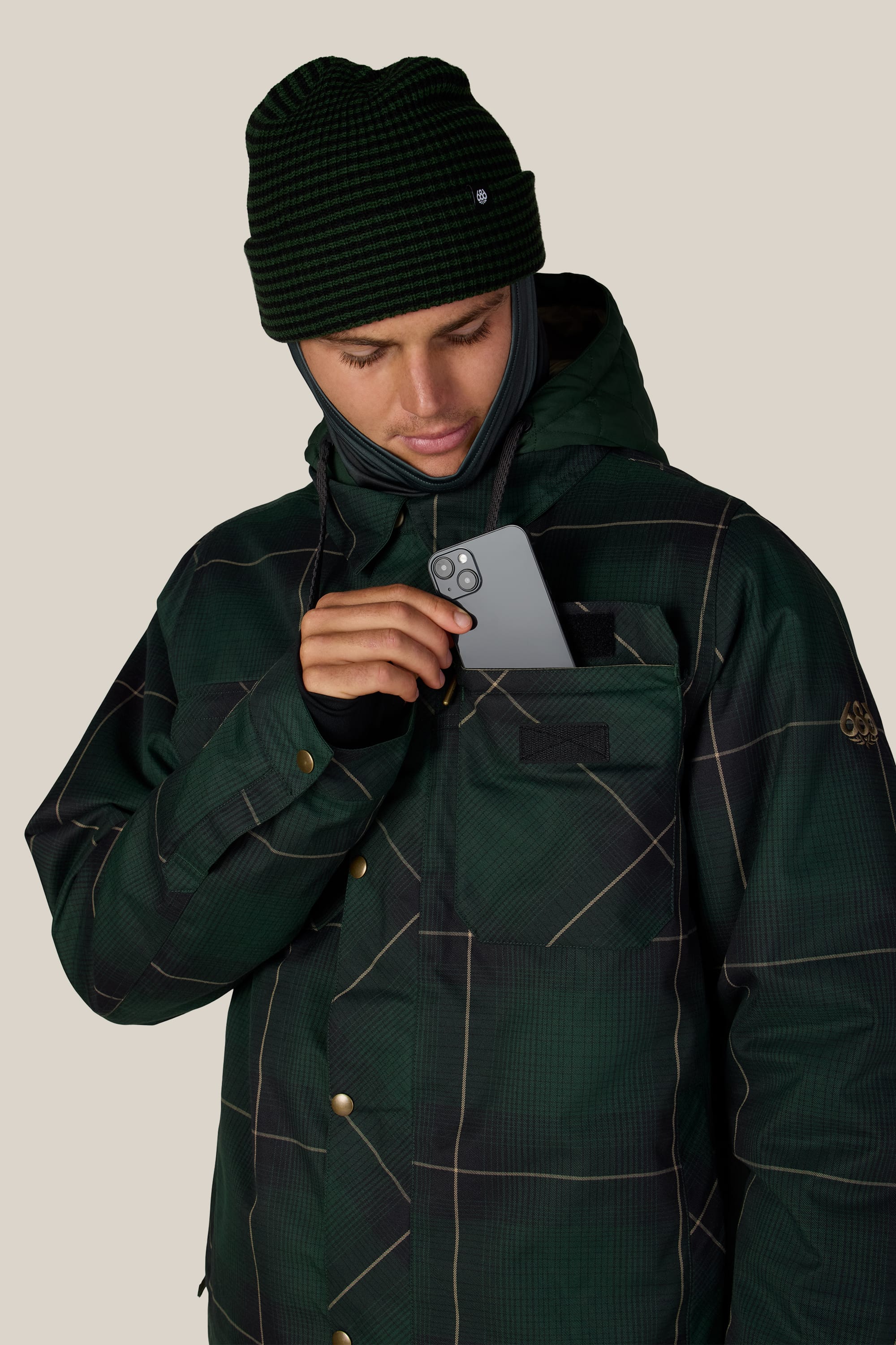 A person wearing the 686 Men’s Woodland™ Insulated Jacket in green plaid and a black beanie places a smartphone into the chest pocket of their jacket, looking down at the phone. Neutral background.