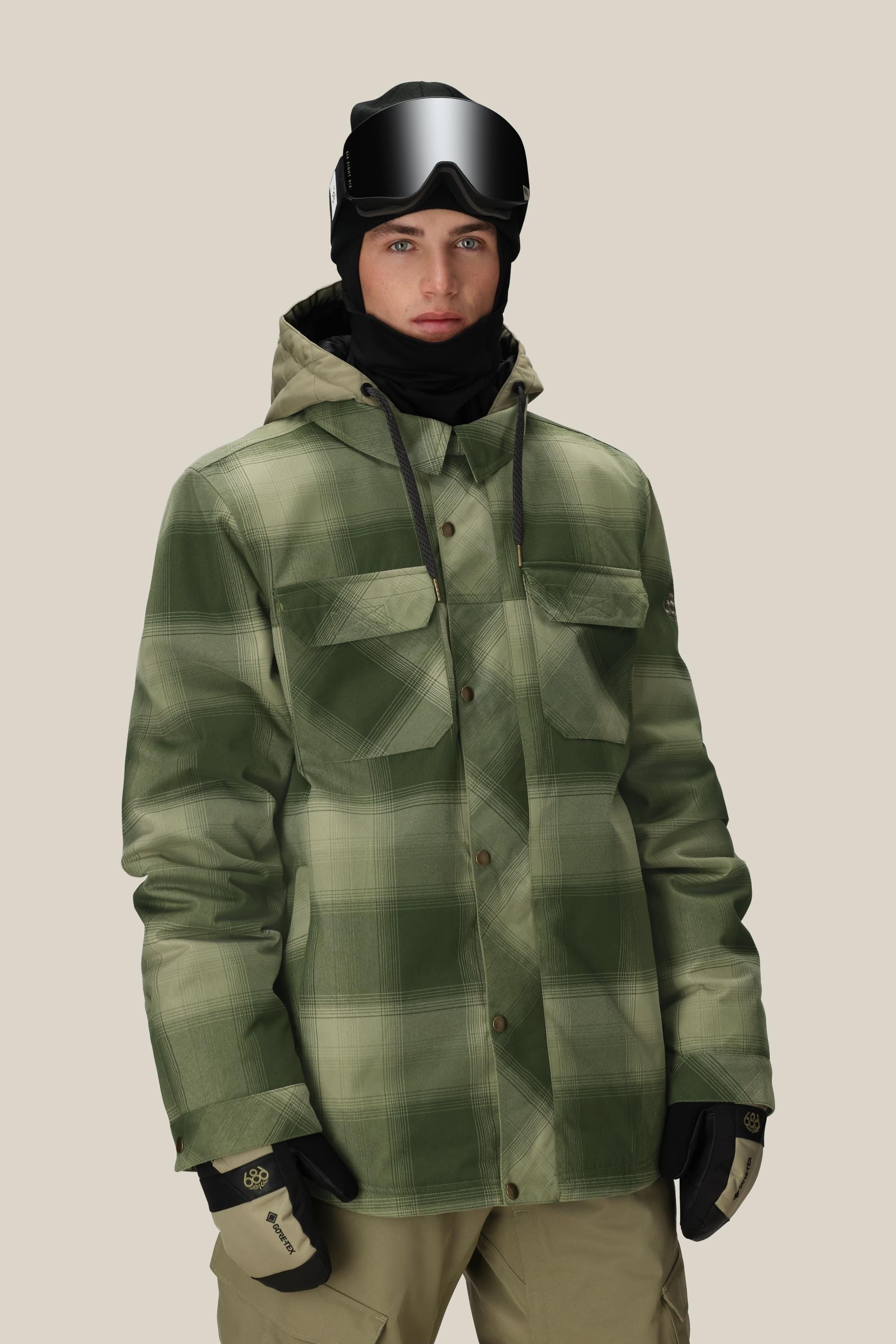 A person wearing the 686 Mens Woodland Insulated Jacket in green plaid, tan snow pants, black gloves, a black ski mask, and ski goggles stands against a plain beige background.