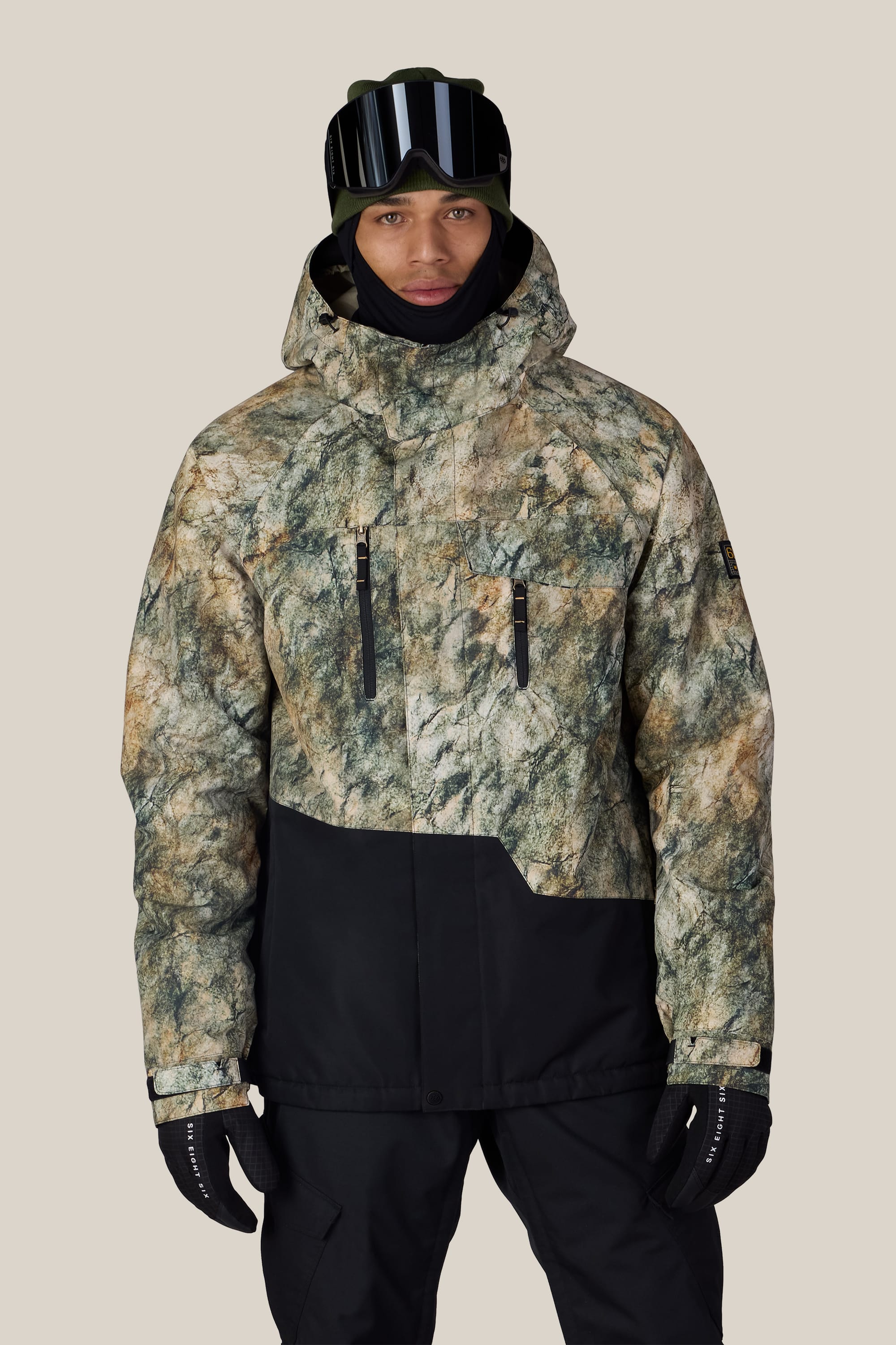 Wearing the 686 Men’s Geo™ Insulated Jacket, a person stands against a plain light background. The jacket features a green and brown camouflage pattern, paired with black snow pants, gloves, ski goggles, and a hood.