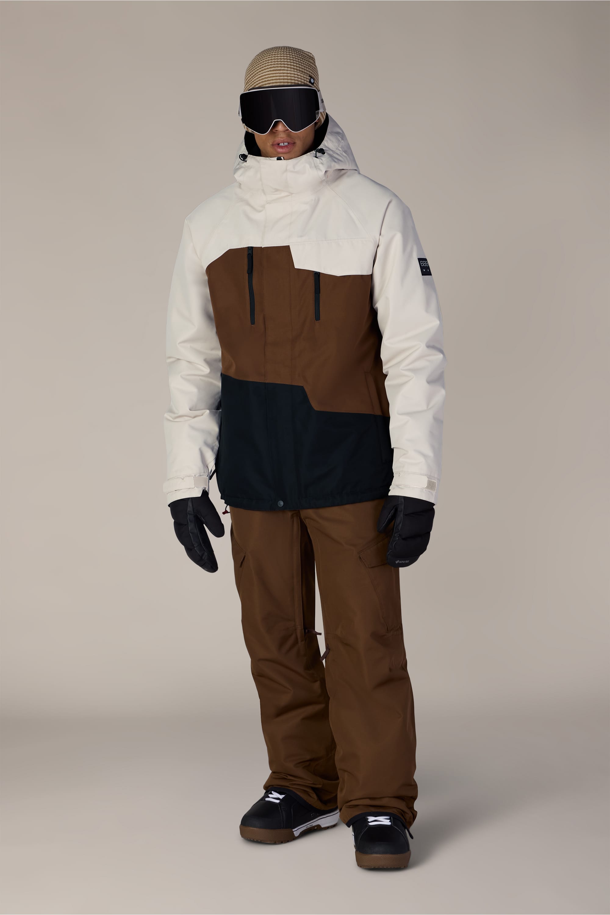 A person wearing the 686 Men’s Geo™ Insulated Jacket in beige and brown with black accents, paired with matching brown snow pants, black gloves, black goggles, a beige beanie, and black shoes, standing against a plain light background.
