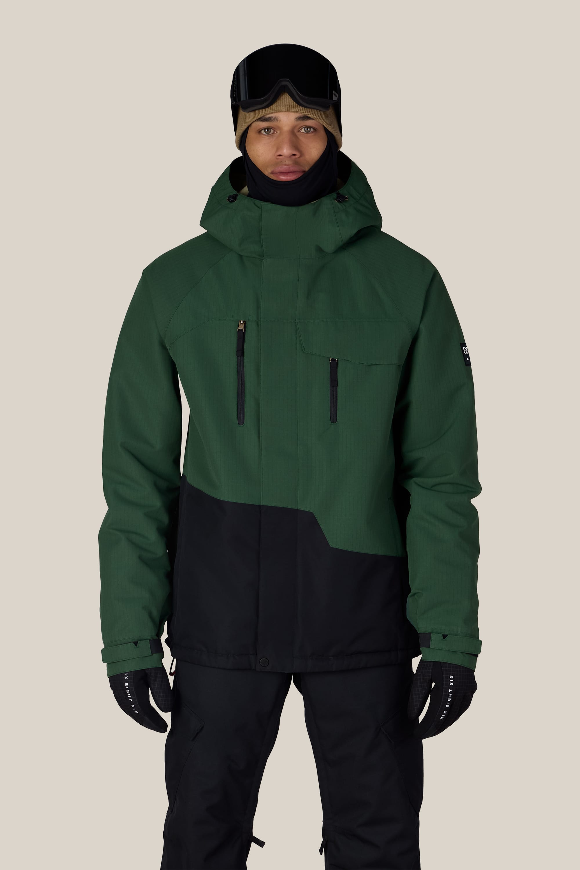 A person wearing the 686 Men’s Geo™ Insulated Jacket from 686 in green and black, paired with black pants, gloves, and ski goggles on their head, standing against a plain light background.