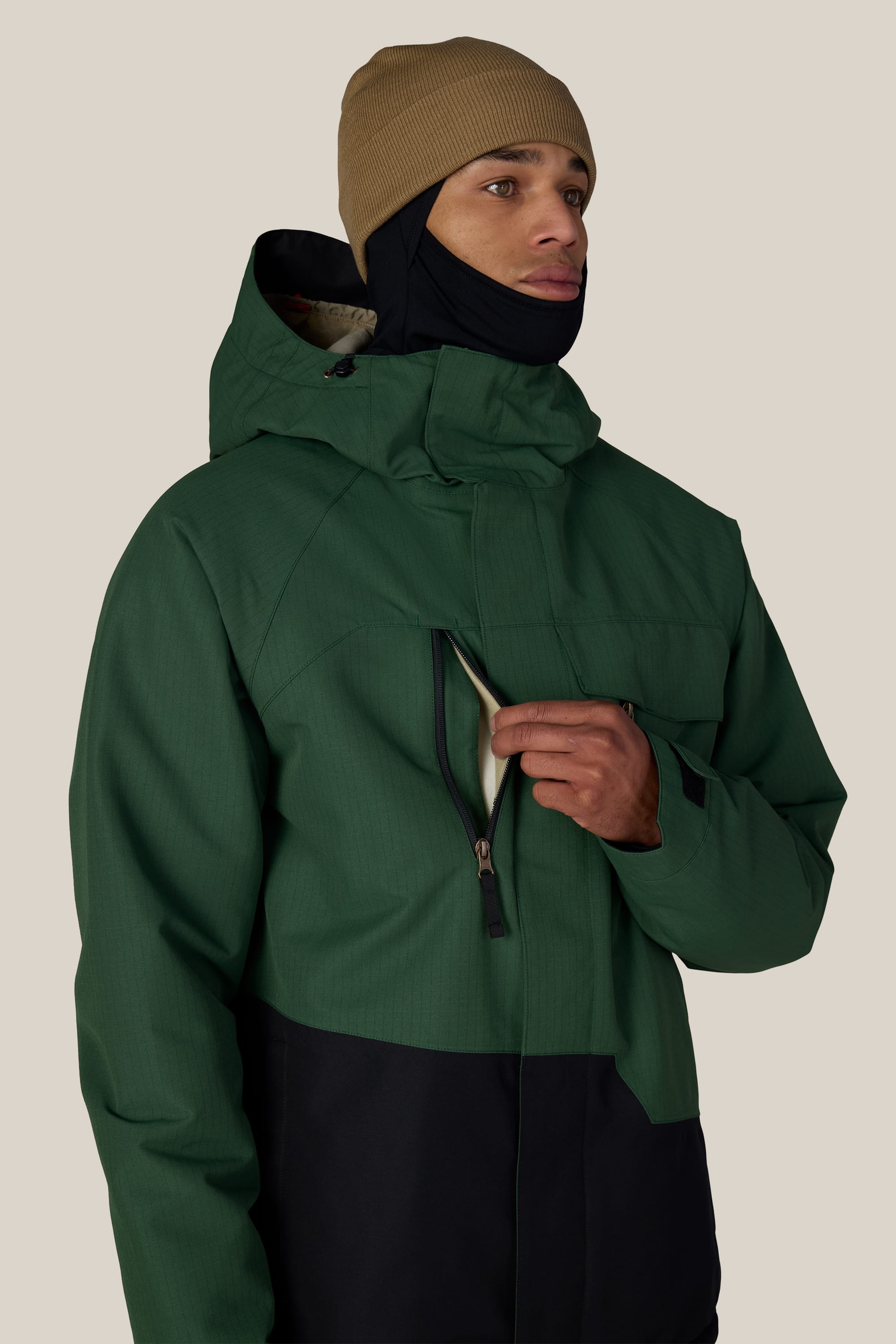 A person wearing the 686 Men’s Geo™ Insulated Jacket in green and black, along with a tan beanie, unzips a chest pocket. The background is plain and light-colored.