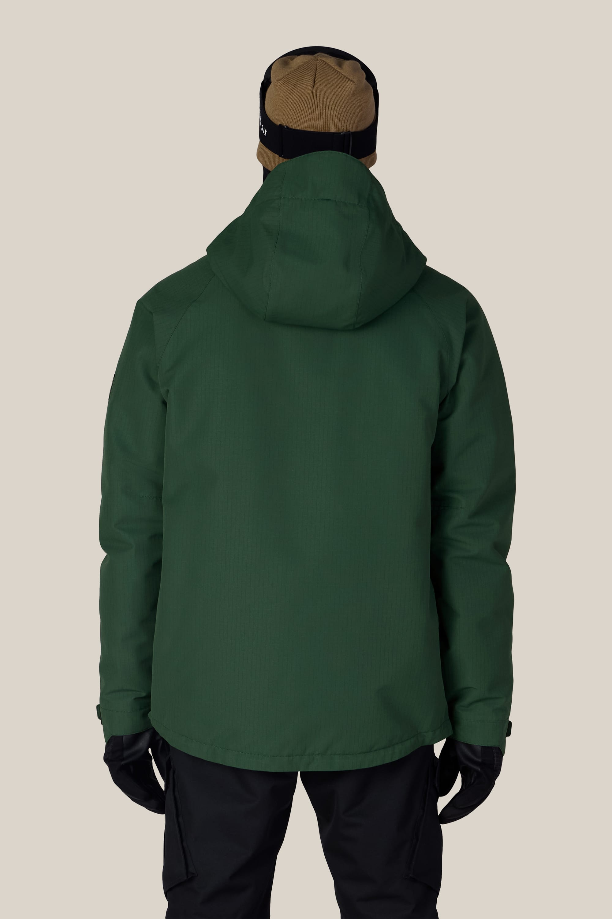 A person wearing the 686 Men’s Geo™ Insulated Jacket in green, paired with black gloves, black pants, and a brown and black beanie, is seen from behind against a plain beige background.