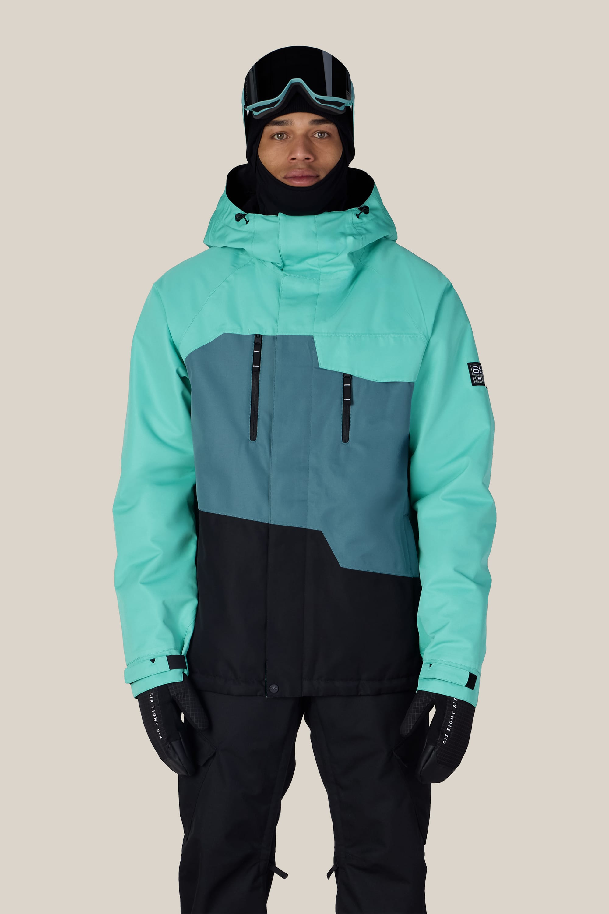 A person wearing the 686 Men’s Geo™ Insulated Jacket in teal, gray, and black from 686, paired with black gloves, ski goggles, and a black hood, stands against a plain light background.