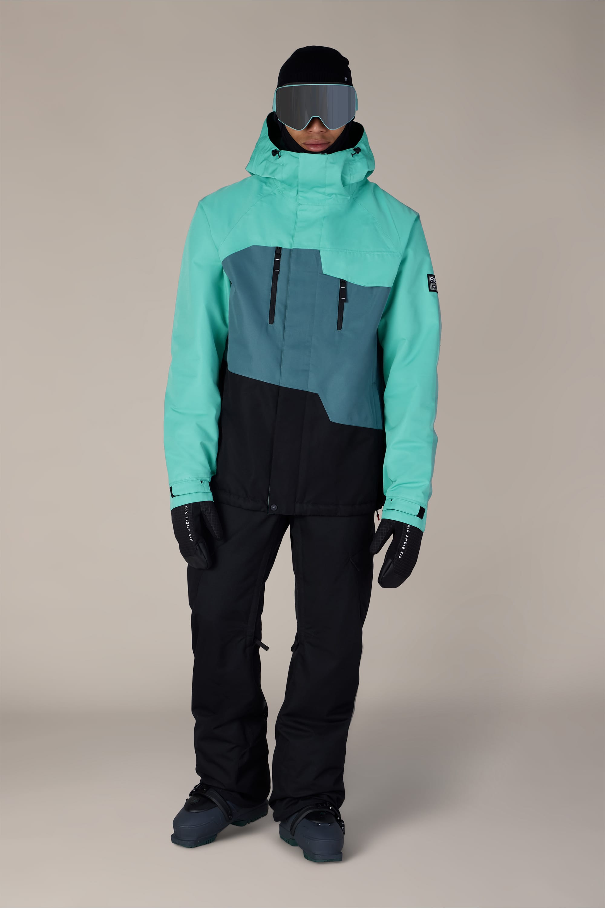 A person wearing a 686 Men’s Geo™ Insulated Jacket in teal and black, along with black snow pants, gloves, boots, ski goggles, and a black beanie stands facing forward against a plain background.