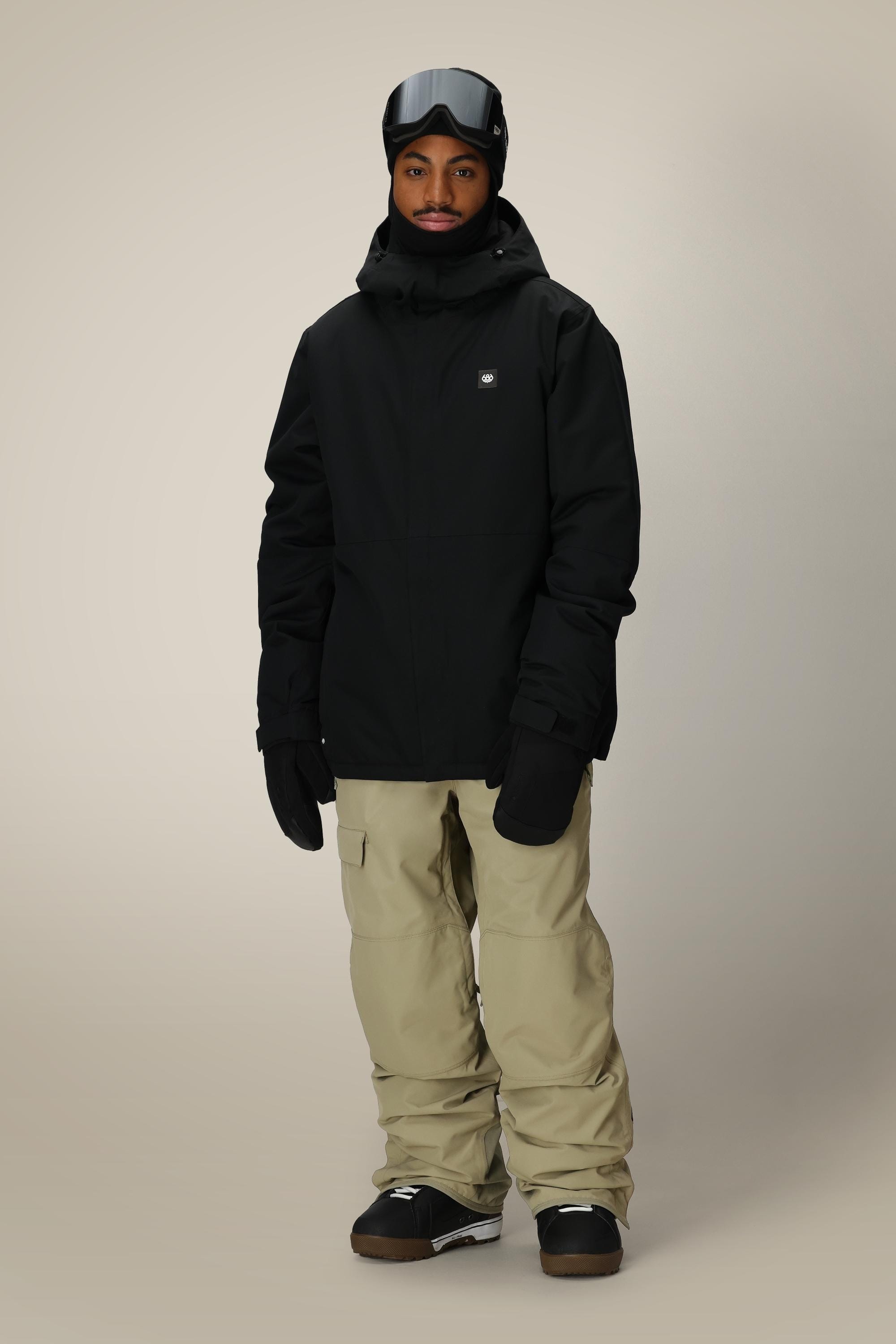 Wearing the 686 Men’s Foundation™ Insulated Jacket in black, along with tan snow pants, black gloves, a black beanie, and ski goggles, a person stands against a plain light background.