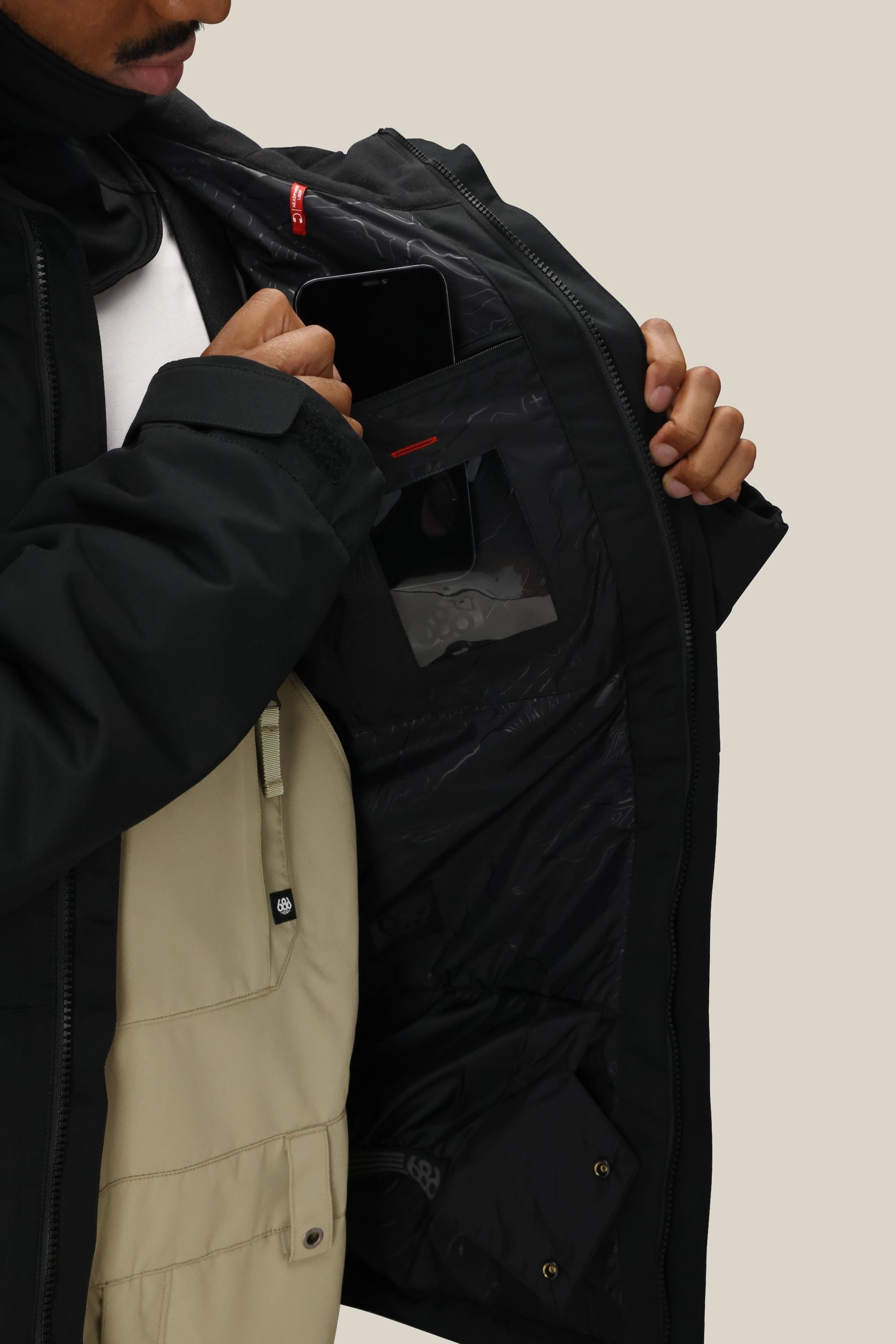 A person opens a black 686 Men’s Foundation™ Insulated Jacket to reveal an inner pocket holding a smartphone. They are wearing a beige overall underneath the jacket, against a plain, light-colored background.
