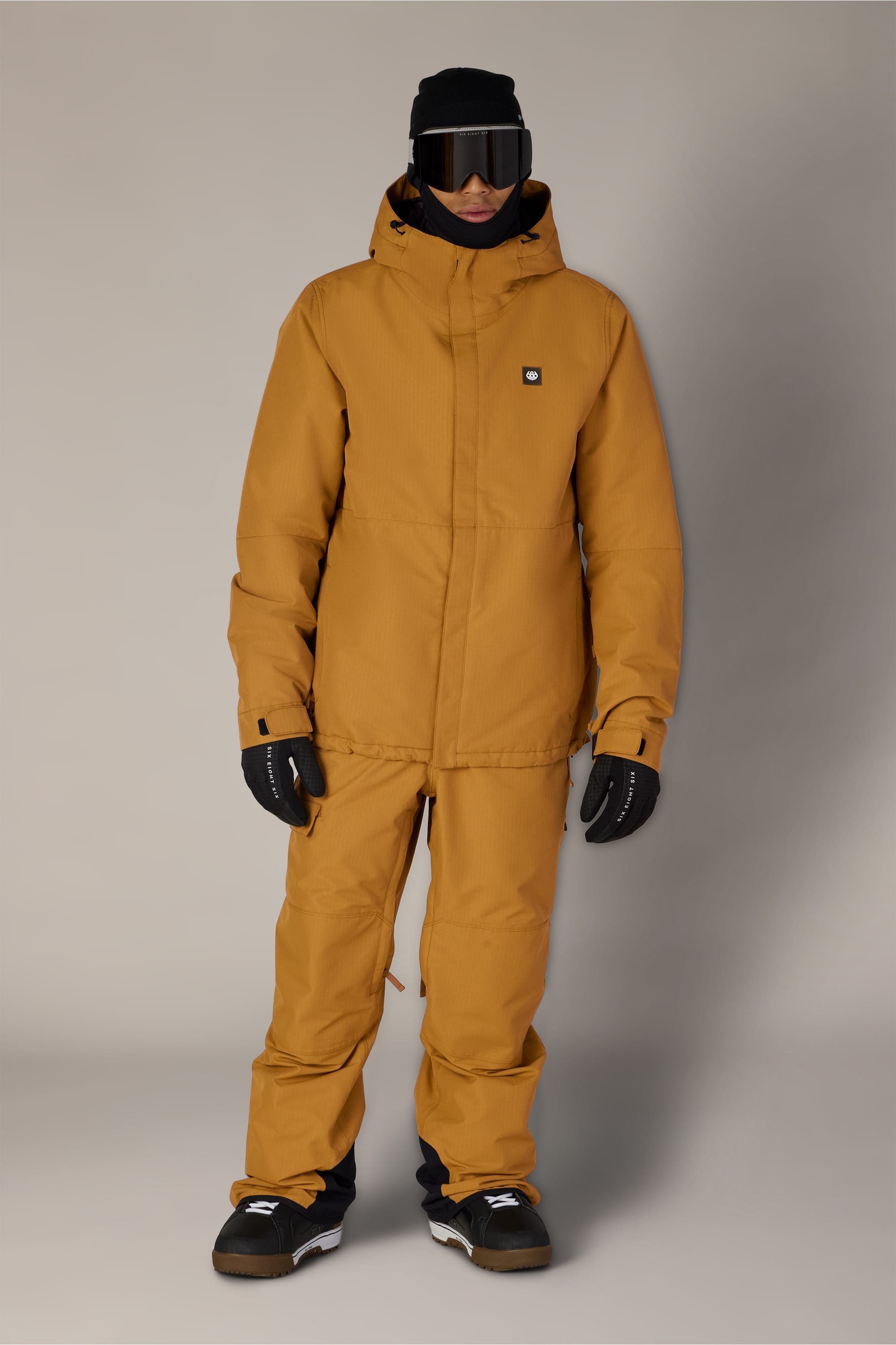 A person stands against a plain, light background wearing the 686 Men’s Foundation™ Insulated Jacket in mustard yellow along with matching pants, black gloves, black ski goggles, a black hat, and black boots.