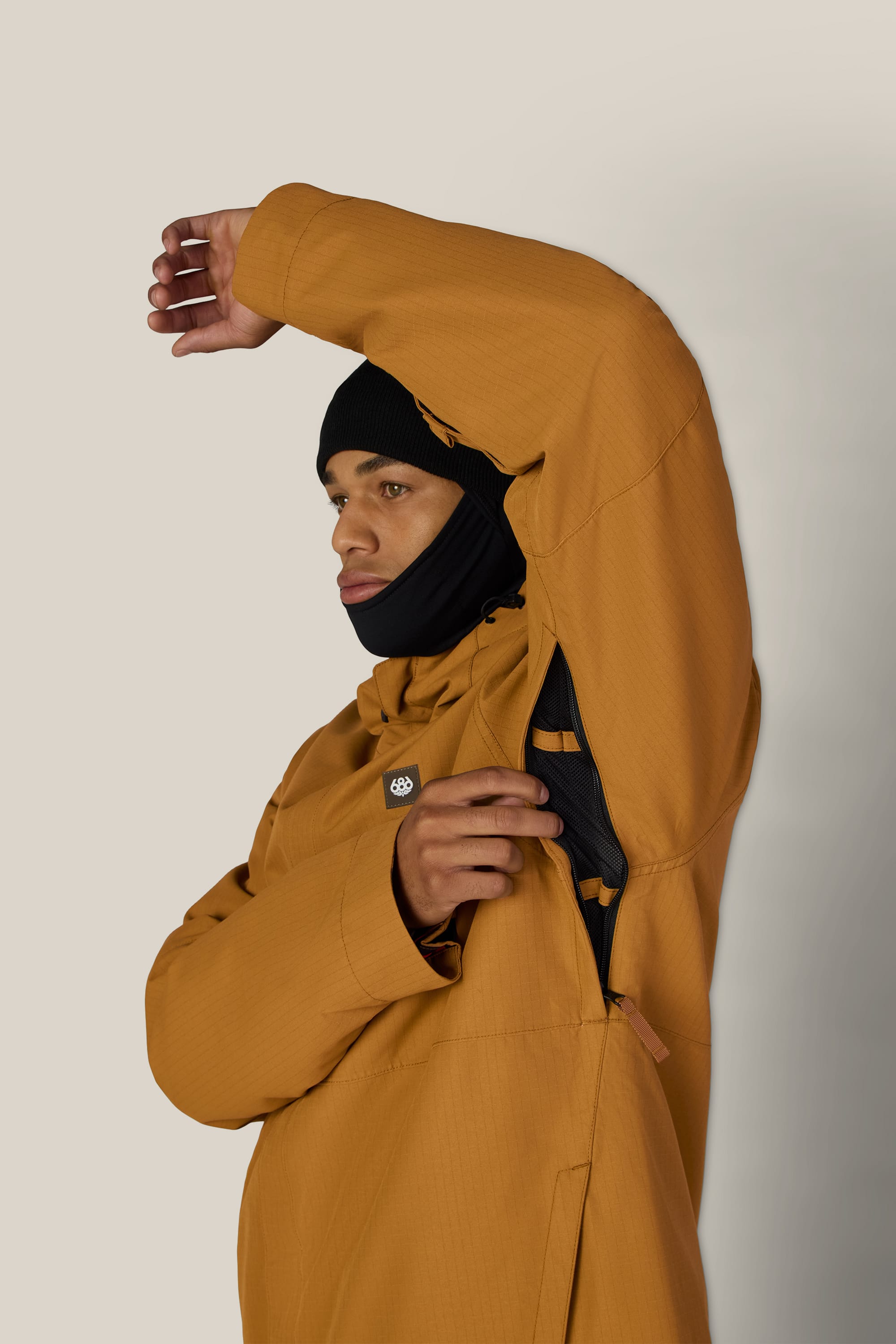 A person wearing the 686 Men’s Foundation™ Insulated Jacket in brown with a black beanie lifts their arm, revealing a zippered underarm vent. The background is plain and light-colored.