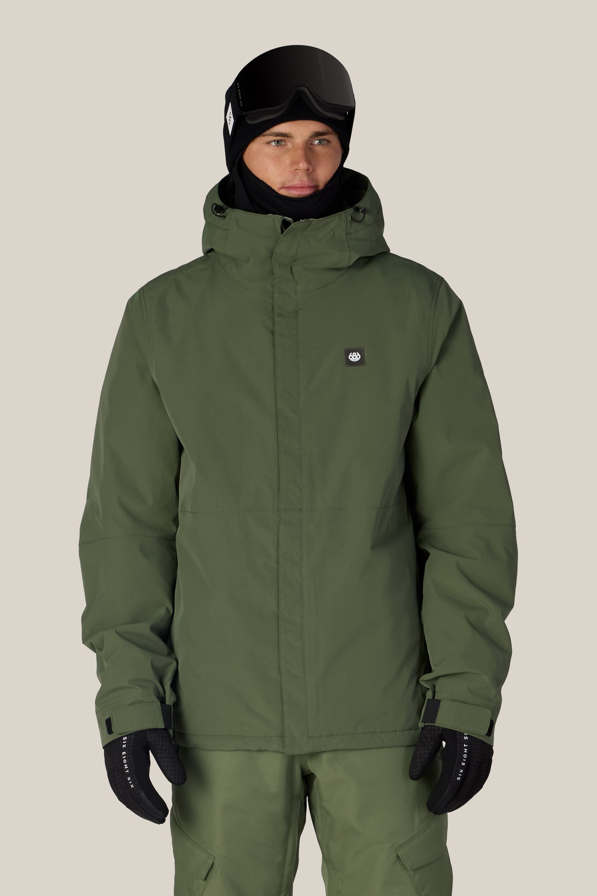 A person wearing the 686 Men’s Foundation™ Insulated Jacket in olive green, paired with matching pants, black gloves, black ski goggles, and a black balaclava, standing against a plain light background.