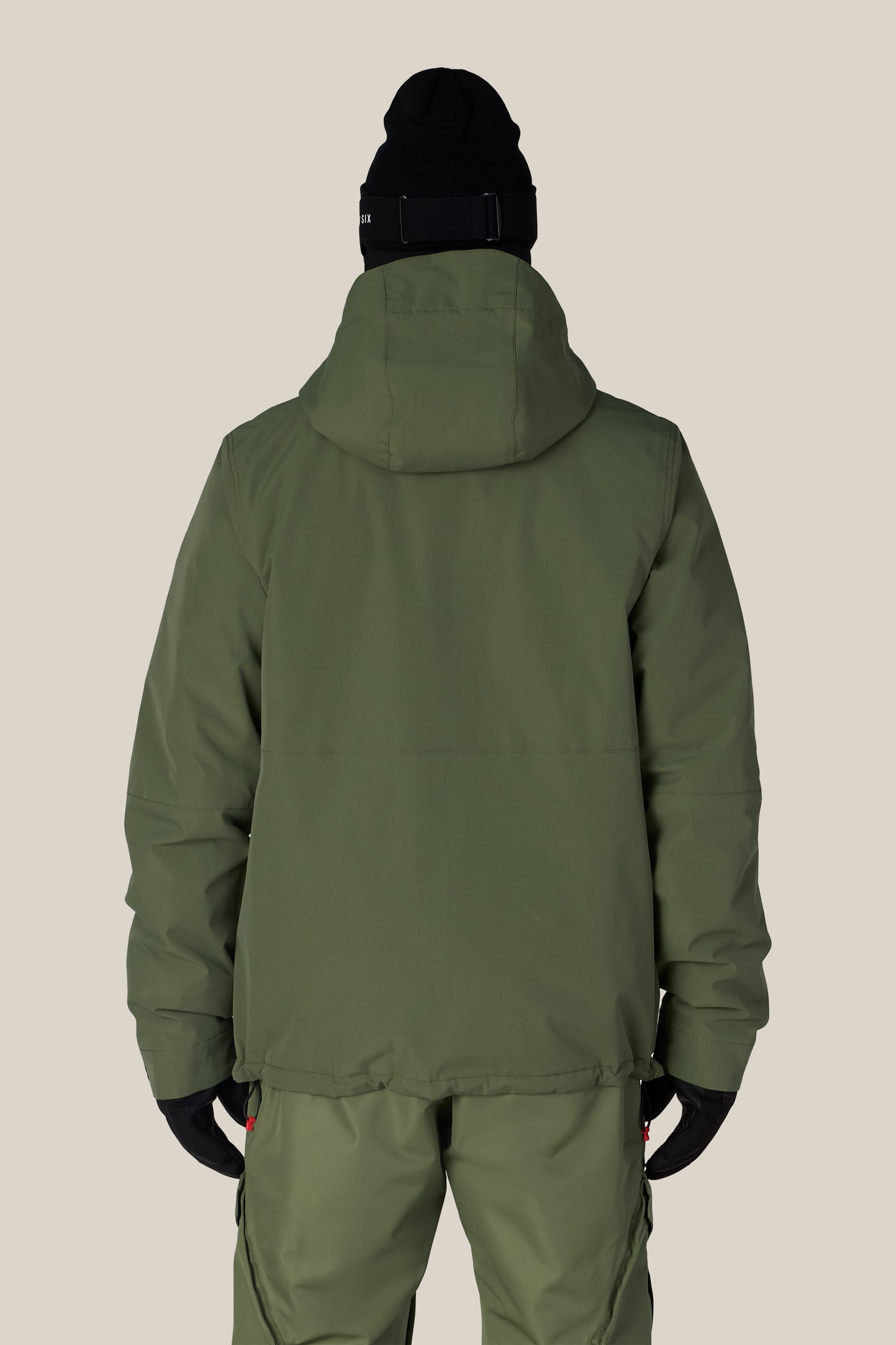 A person wearing the 686 Men’s Foundation™ Insulated Jacket in olive green with a hood, paired with matching pants, black gloves, and a black beanie stands facing away from the camera against a plain beige background.