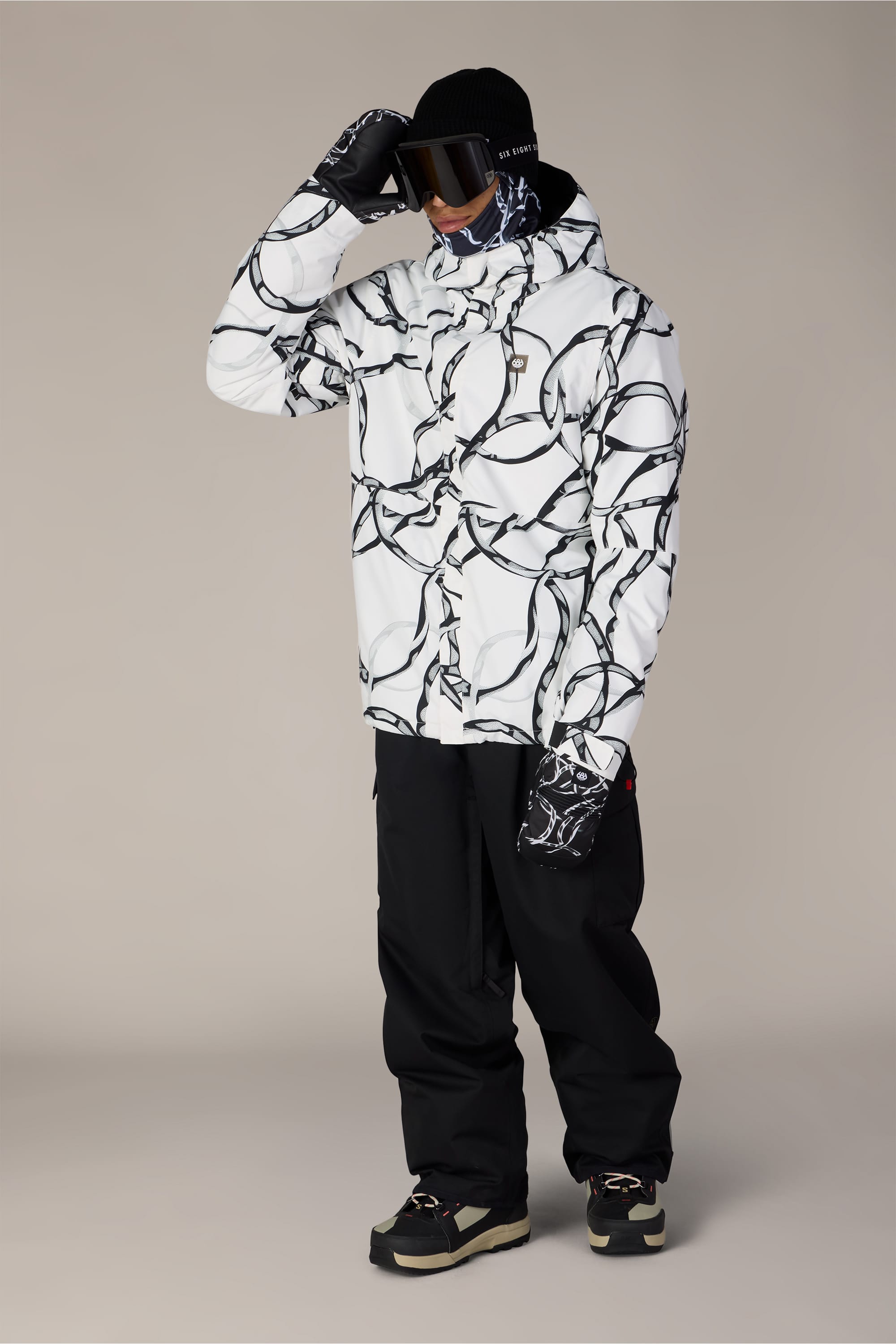 A person wearing the 686 Men’s Foundation™ Insulated Jacket by 686—which features a white color with a black abstract pattern—paired with black snow pants, dark gloves, a black hat, face covering, and ski goggles, standing against a plain beige background.