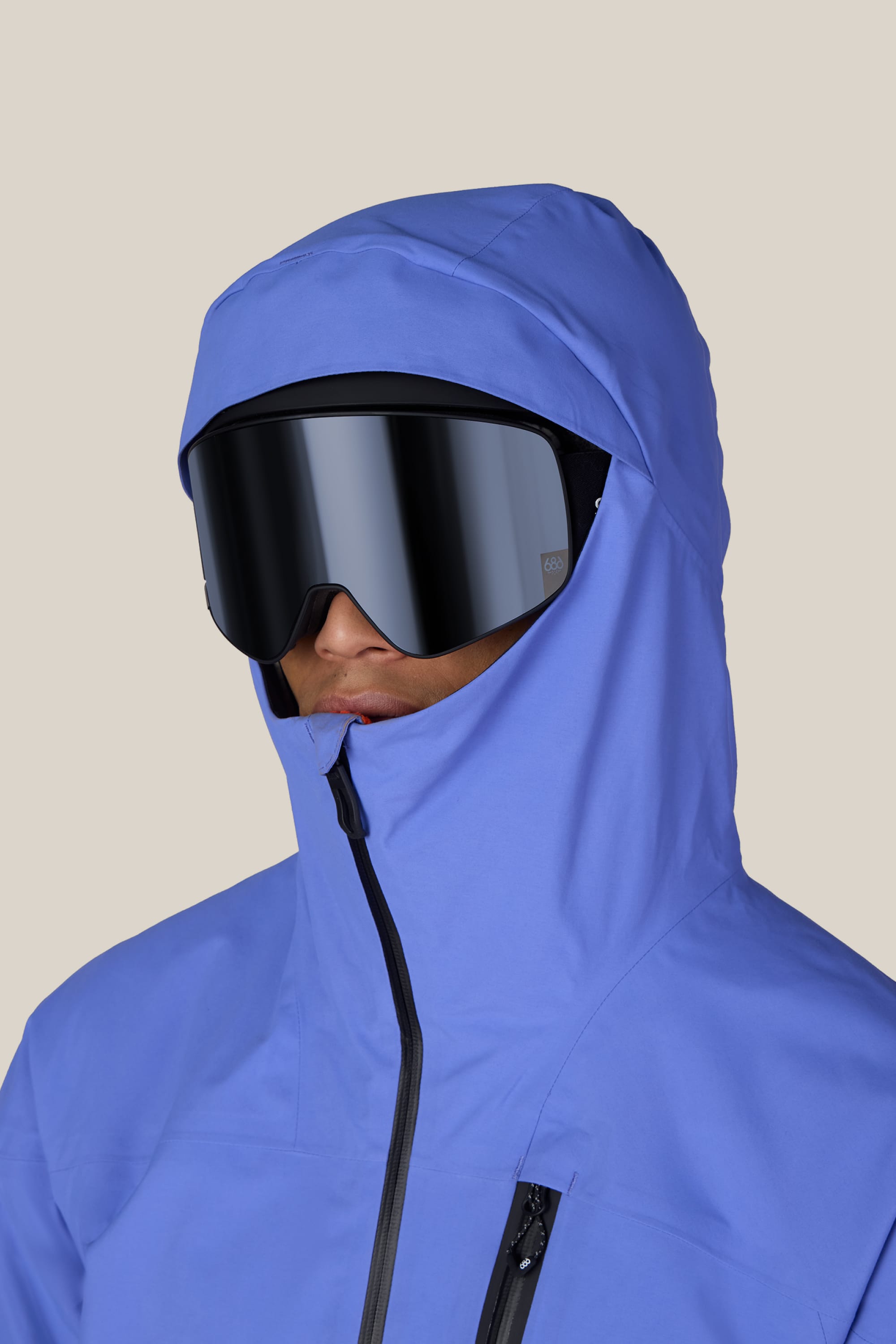 A person wearing the 686 Men’s GORE-TEX® GT Thermagraph® Jacket in blue, featuring a hood and zipper, along with large black ski goggles, is looking forward against a plain light-colored background.