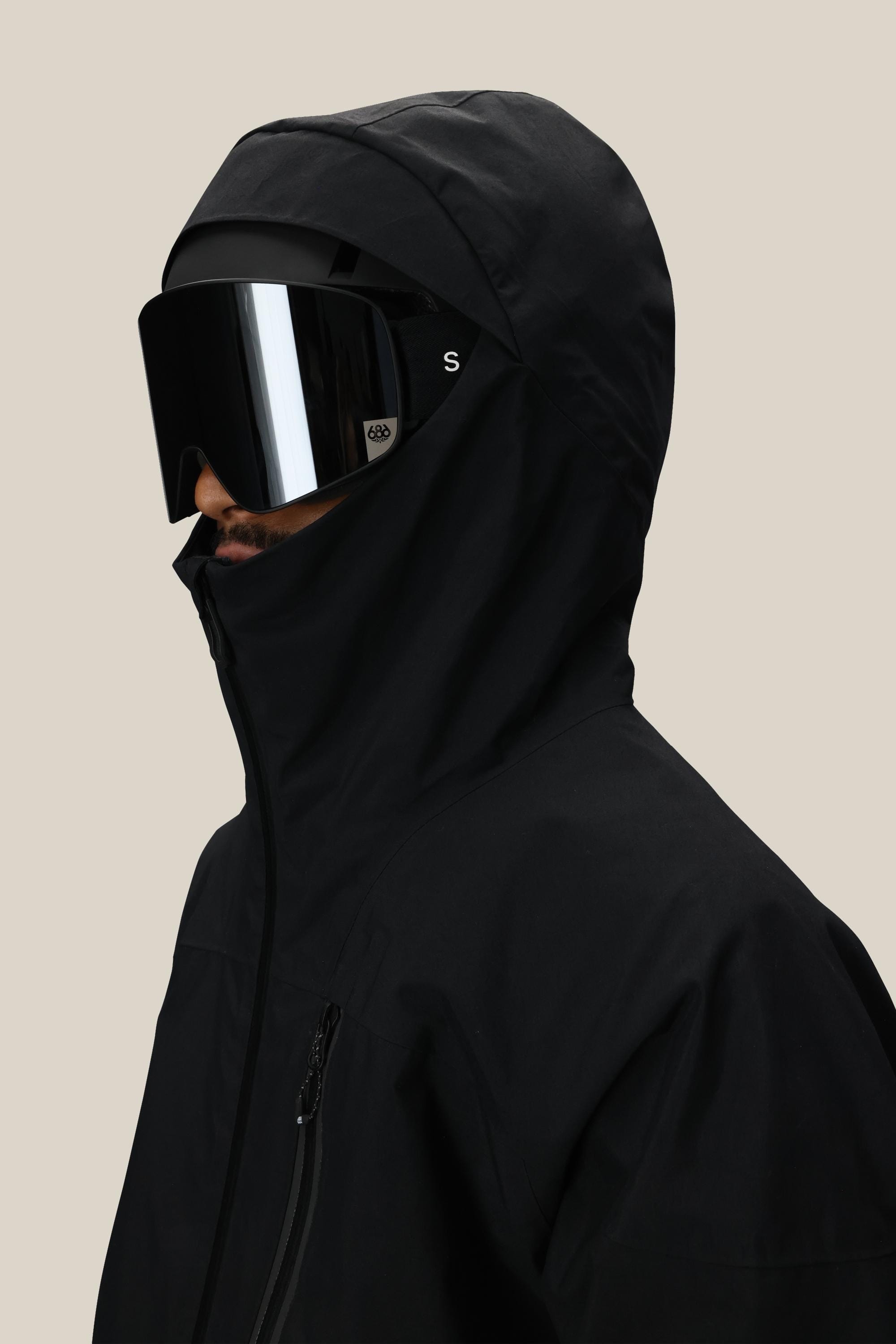 A person wearing the 686 Men’s GORE-TEX® GT Thermagraph® Jacket and large black reflective ski goggles faces left against a beige background.
