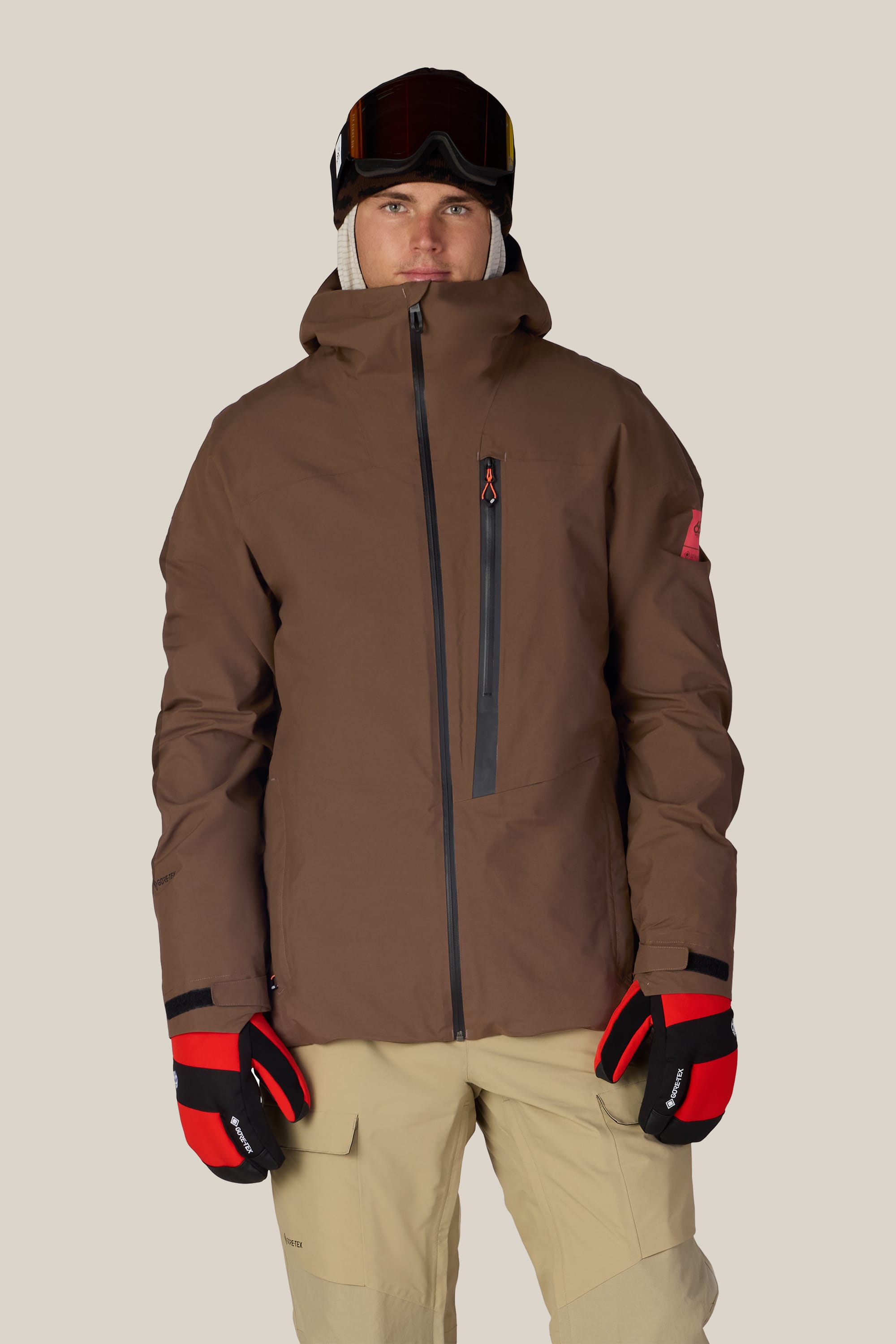 A person wearing the 686 Men’s GORE-TEX® GT Thermagraph® Jacket by 686, beige pants, red and black gloves, ski goggles, and a black helmet stands facing the camera against a plain background.