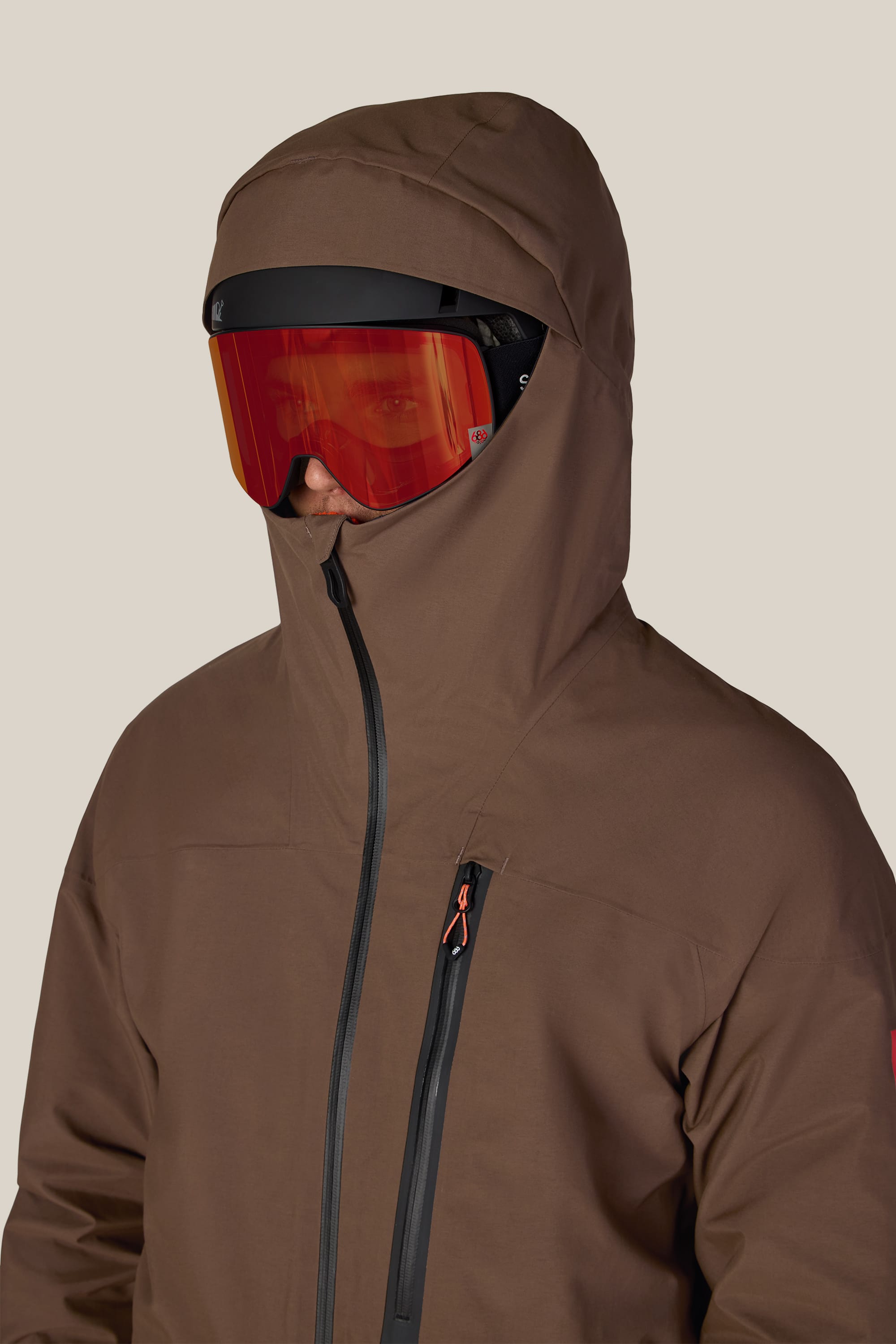 A person wearing the 686 Men’s GORE-TEX® GT Thermagraph® Jacket in brown, fully zipped up to their nose, and sporting large red-orange ski goggles, stands against a plain light background.