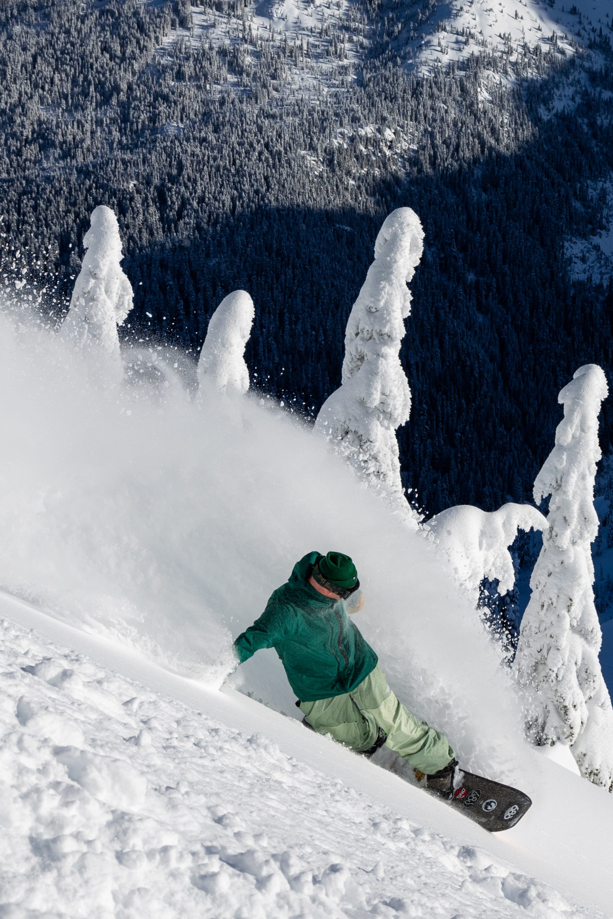 A snowboarder wearing the 686 Mens GORE-TEX GT Thermagraph® Jacket carves through deep powder on a snowy mountain slope, with snow-covered trees and a forested peak in the background.
