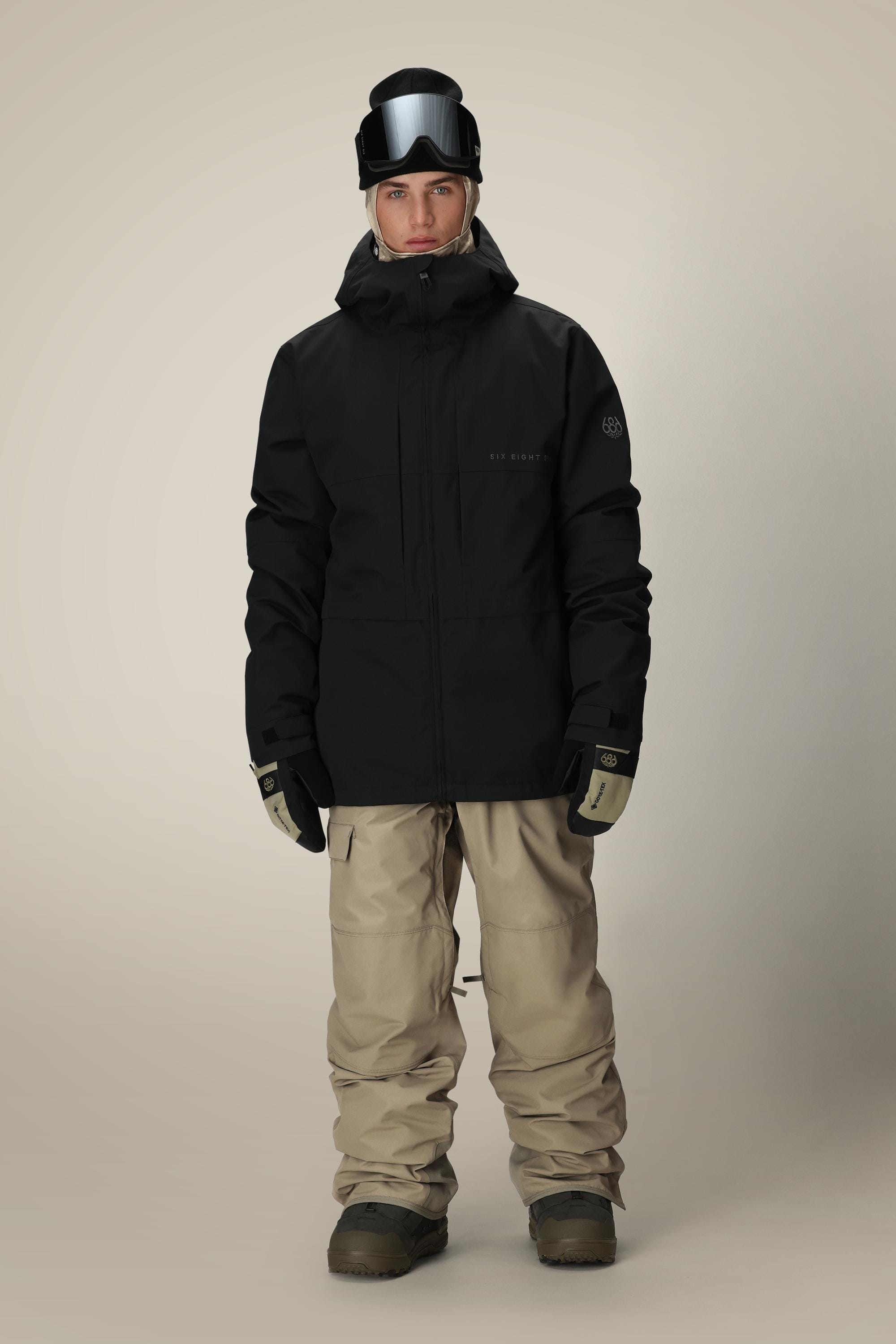 A person wearing the 686 Men’s SMARTY® 3-In-1 Form Jacket in black, along with beige snow pants, gloves, boots, and black ski goggles on their head, stands facing forward against a plain background.