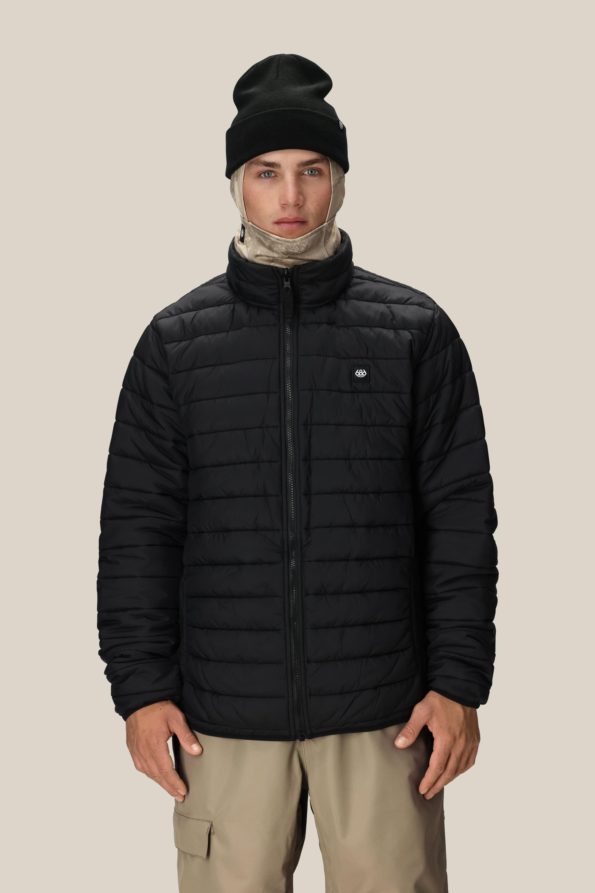 A person wearing the 686 Men’s SMARTY® 3-In-1 Form Jacket in black, tan pants, a beige balaclava, and a black beanie stands against a plain light background, facing forward with hands relaxed by their sides.