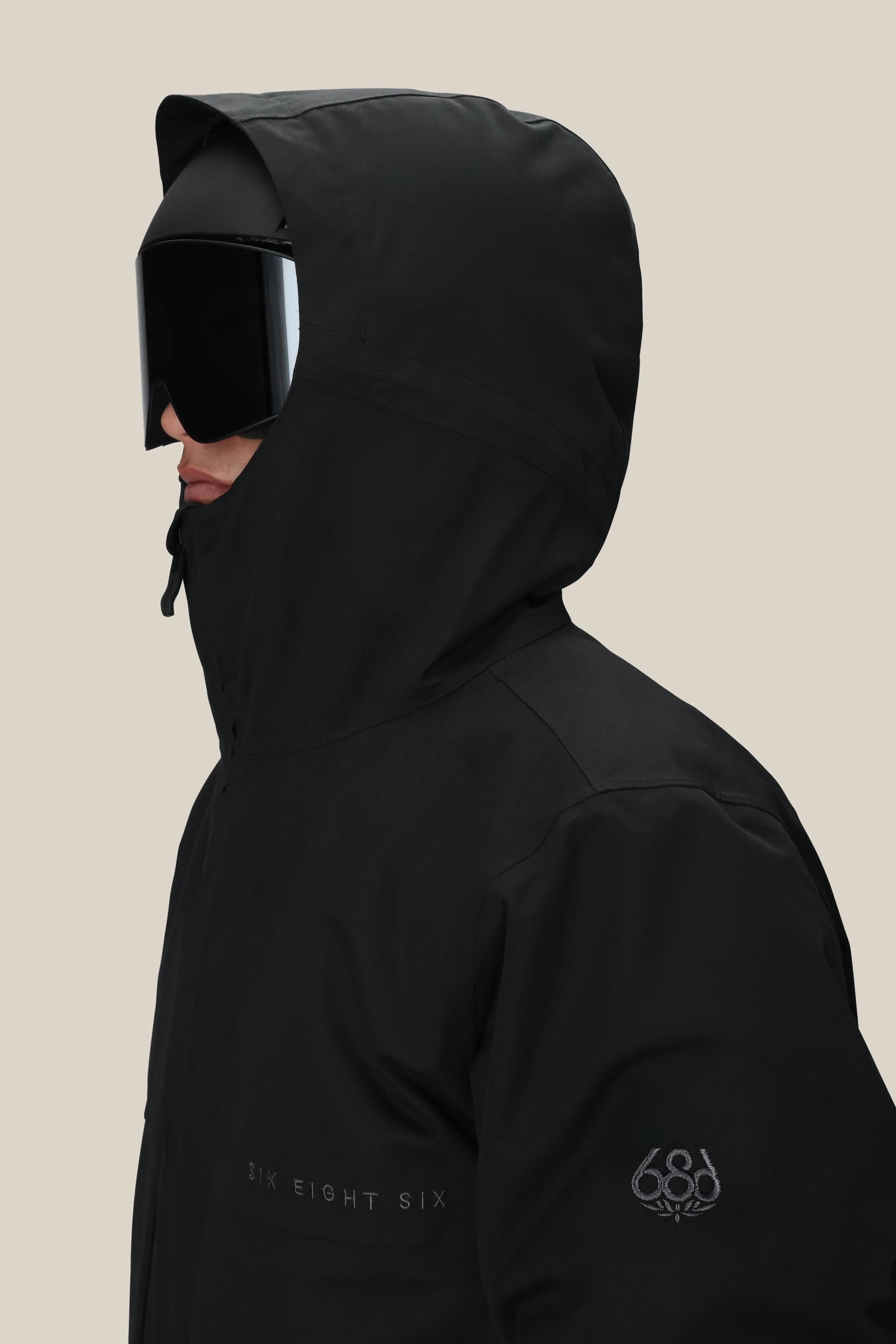 A person wearing the 686 Men’s SMARTY® 3-In-1 Form Jacket in black, featuring the Six Eight Six branding printed on the chest and a 686 logo on the sleeve, is shown from the side against a plain beige background. The individual is also wearing large black goggles and a hood over their head.