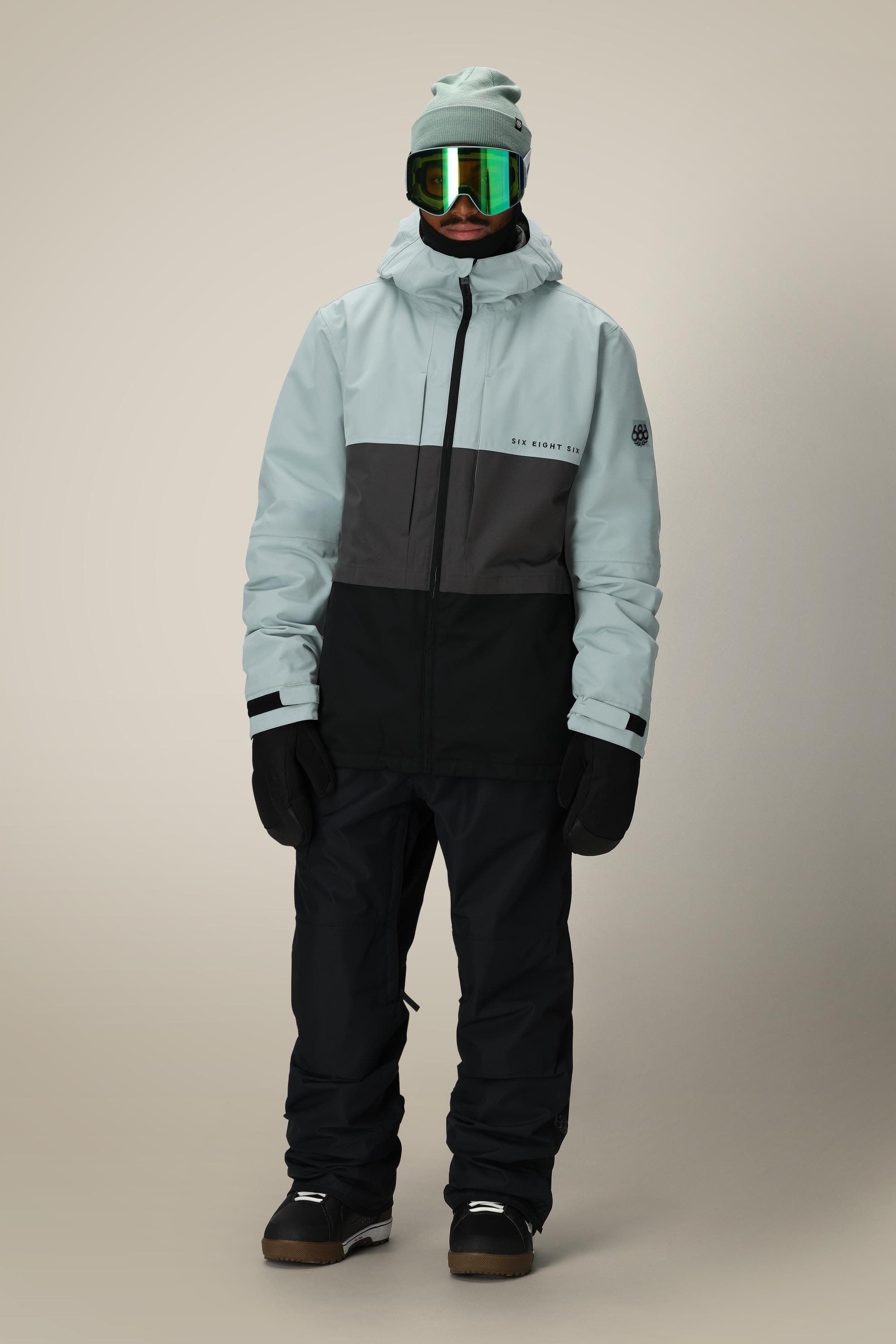 A person wearing the 686 Mens Smarty 3-In-1 Form Jacket in light blue and black, paired with black snow pants, gloves, boots, green ski goggles, and a beanie stands against a plain background.