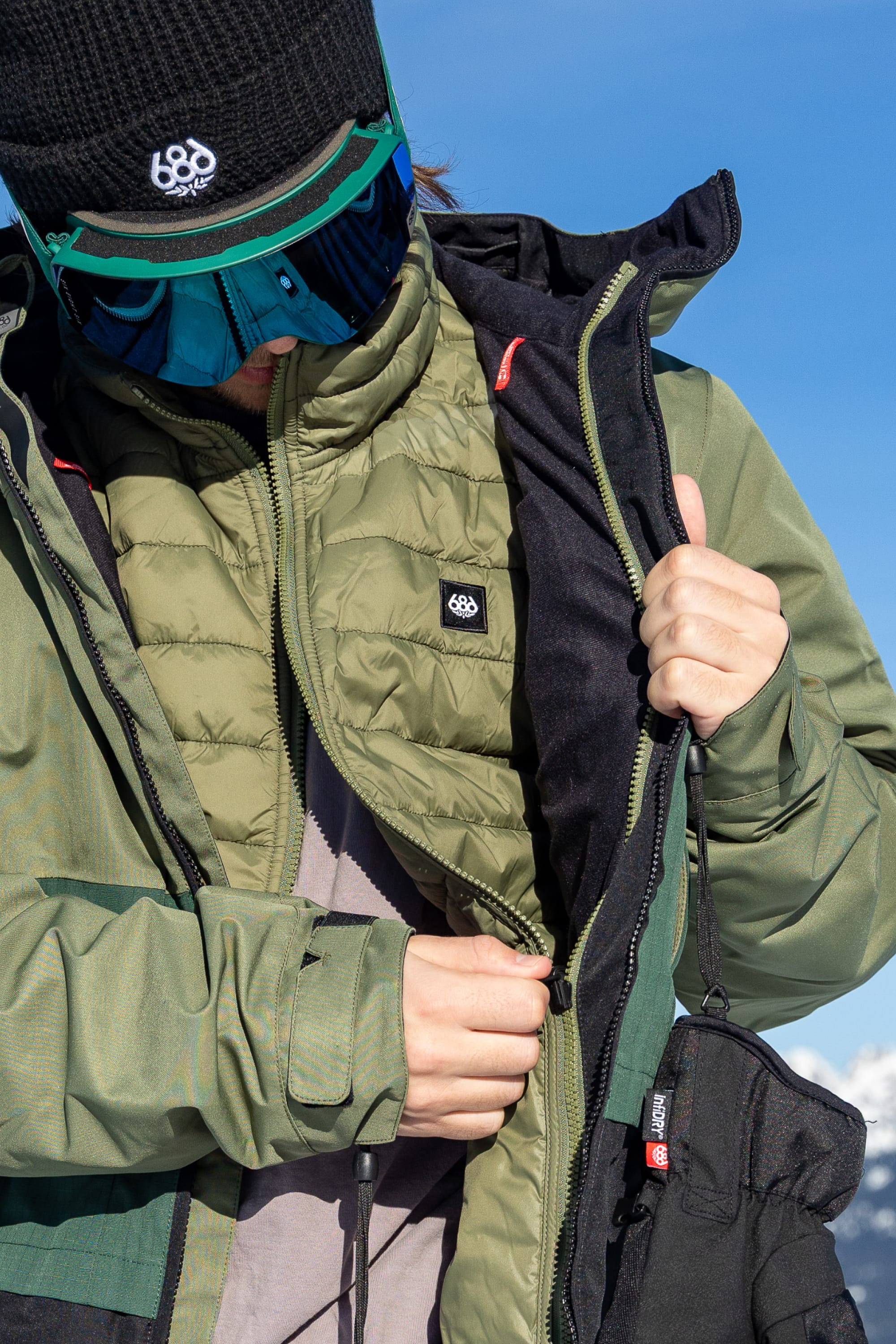 A person dressed in green and black winter gear zips up the 686 Men’s SMARTY® 3-In-1 Form Jacket, complemented by a black beanie, reflective ski goggles, and a green quilted vest, with snow and blue sky in the background.