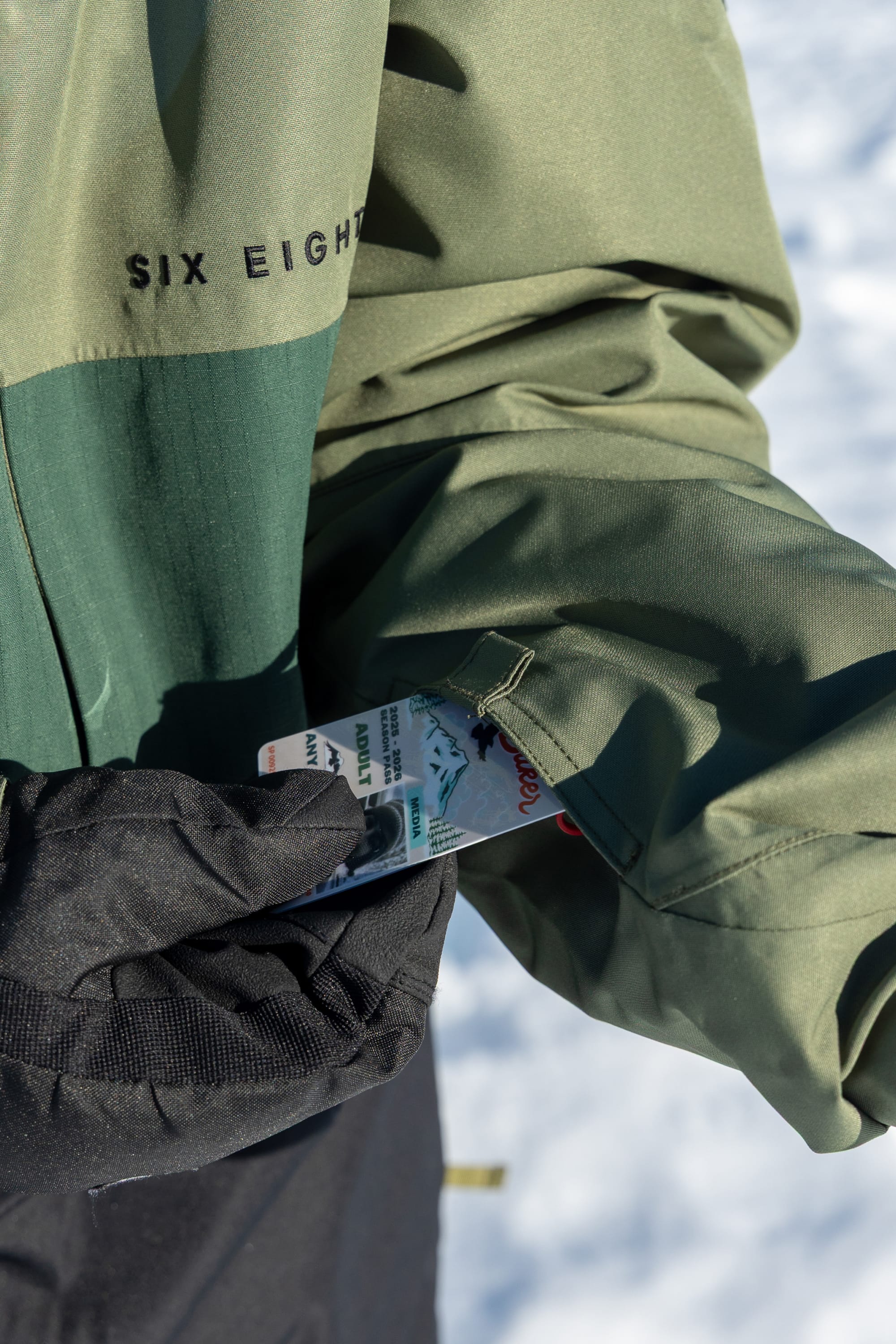 Wearing the 686 Men’s SMARTY® 3-In-1 Form Jacket, a person pulls ski lift passes from a sleeve pocket, with snow in the background.
