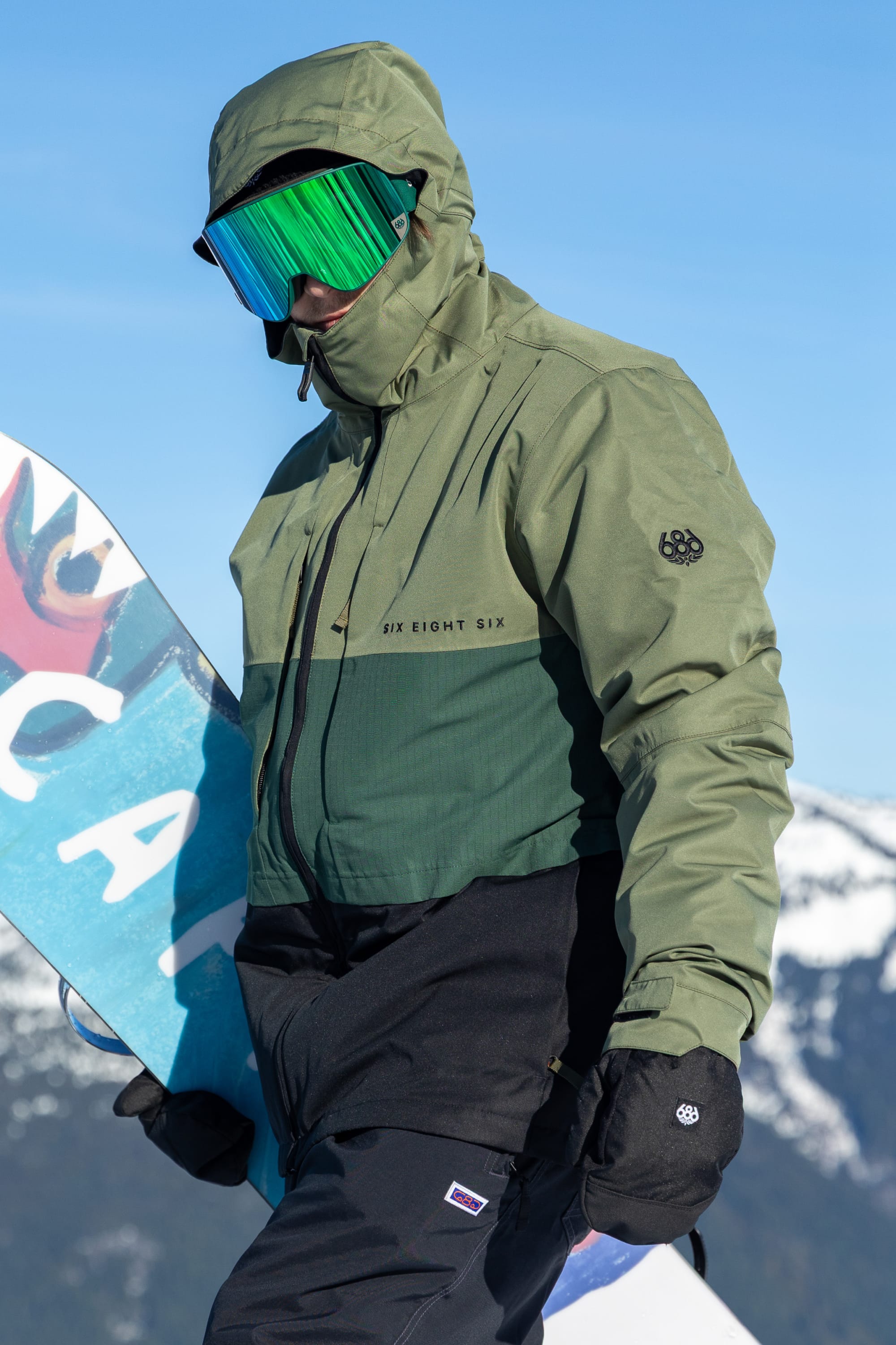 A person wearing the 686 Men’s SMARTY® 3-In-1 Form Jacket by 686, along with black gloves and reflective ski goggles, holds a snowboard on a snowy mountain under a clear blue sky.