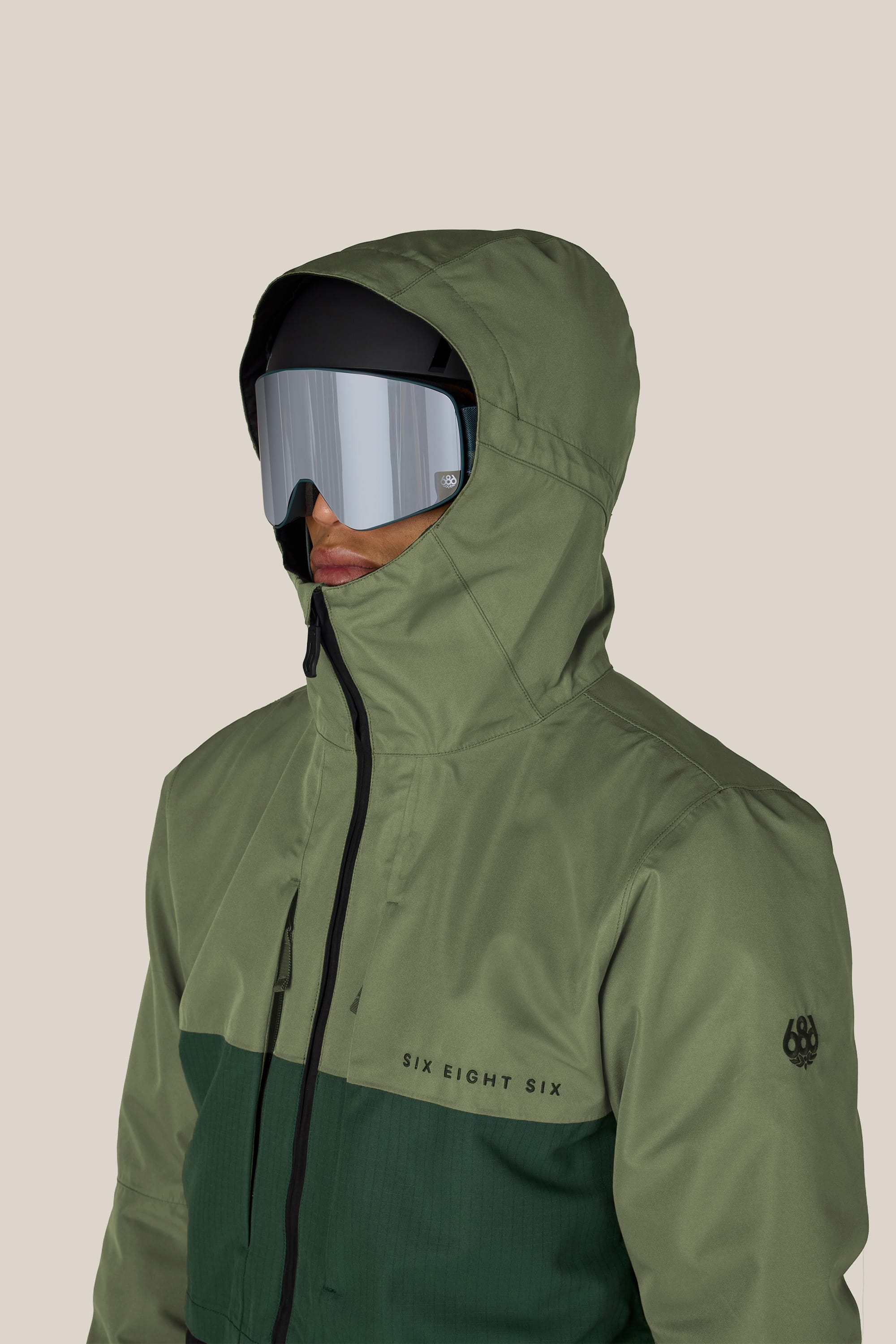 A person is wearing the 686 Men’s SMARTY® 3-In-1 Form Jacket in green, paired with reflective ski goggles and a black base layer. The jacket features the 686 brand name on the chest and a logo on the sleeve, set against a beige background.