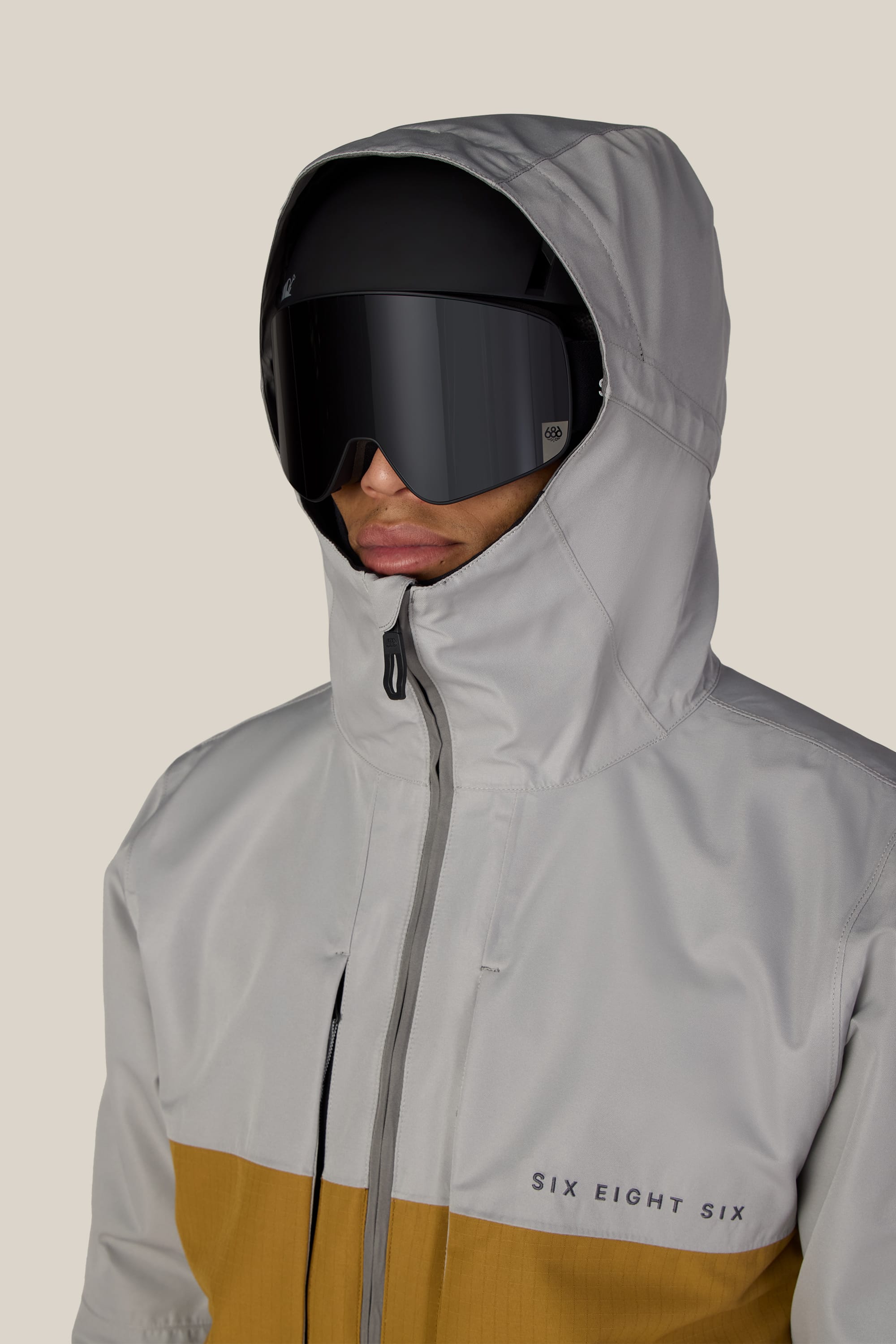 A person wearing the 686 Men’s SMARTY® 3-In-1 Form Jacket in gray and tan with prominent 686 branding, along with large black ski goggles, is facing forward against a plain light background.