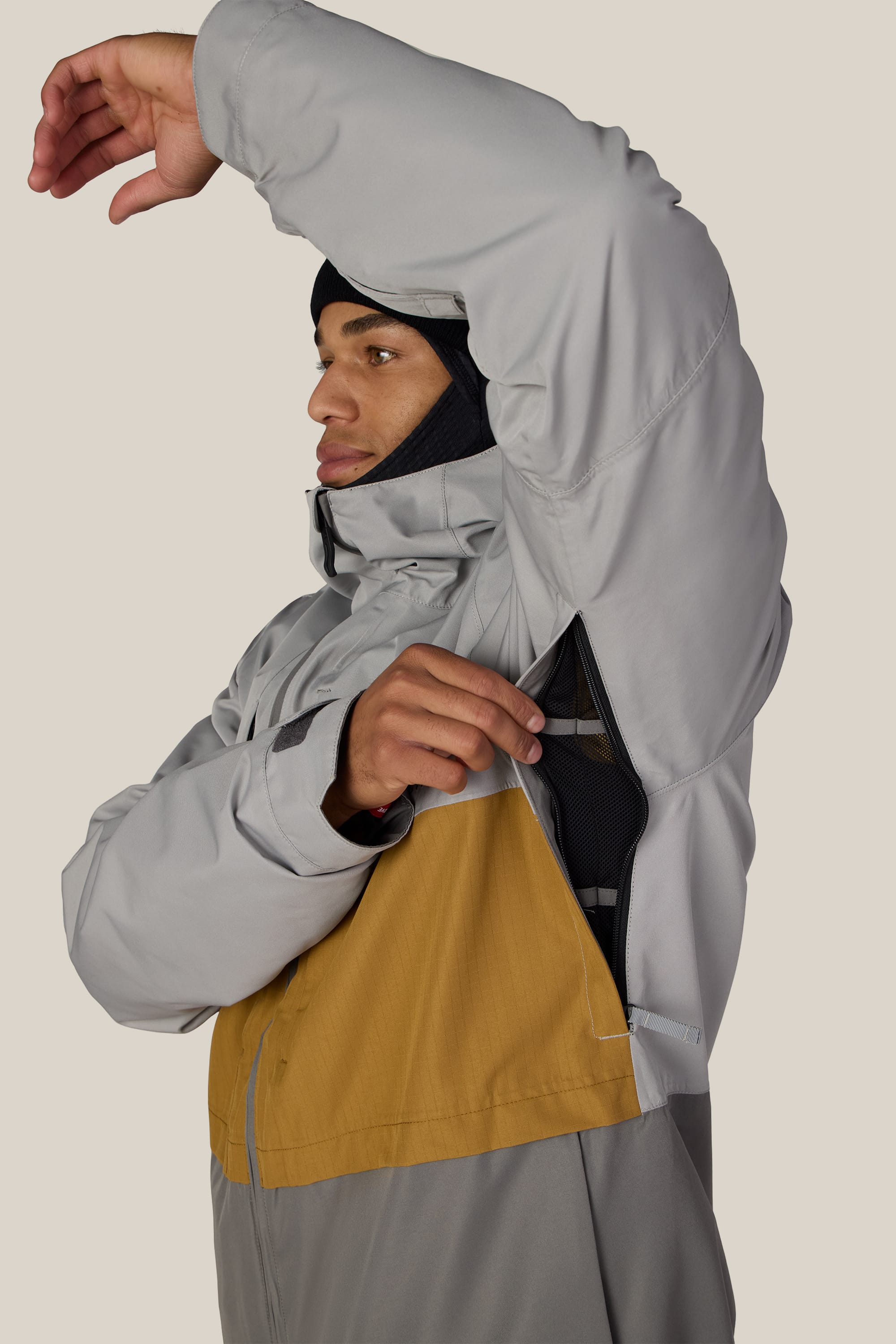 A person wearing the 686 Men’s SMARTY® 3-In-1 Form Jacket in gray and yellow lifts one arm to unzip a ventilation opening under the sleeve. Set against a plain beige background, they also wear a black hood or balaclava beneath the jacket.