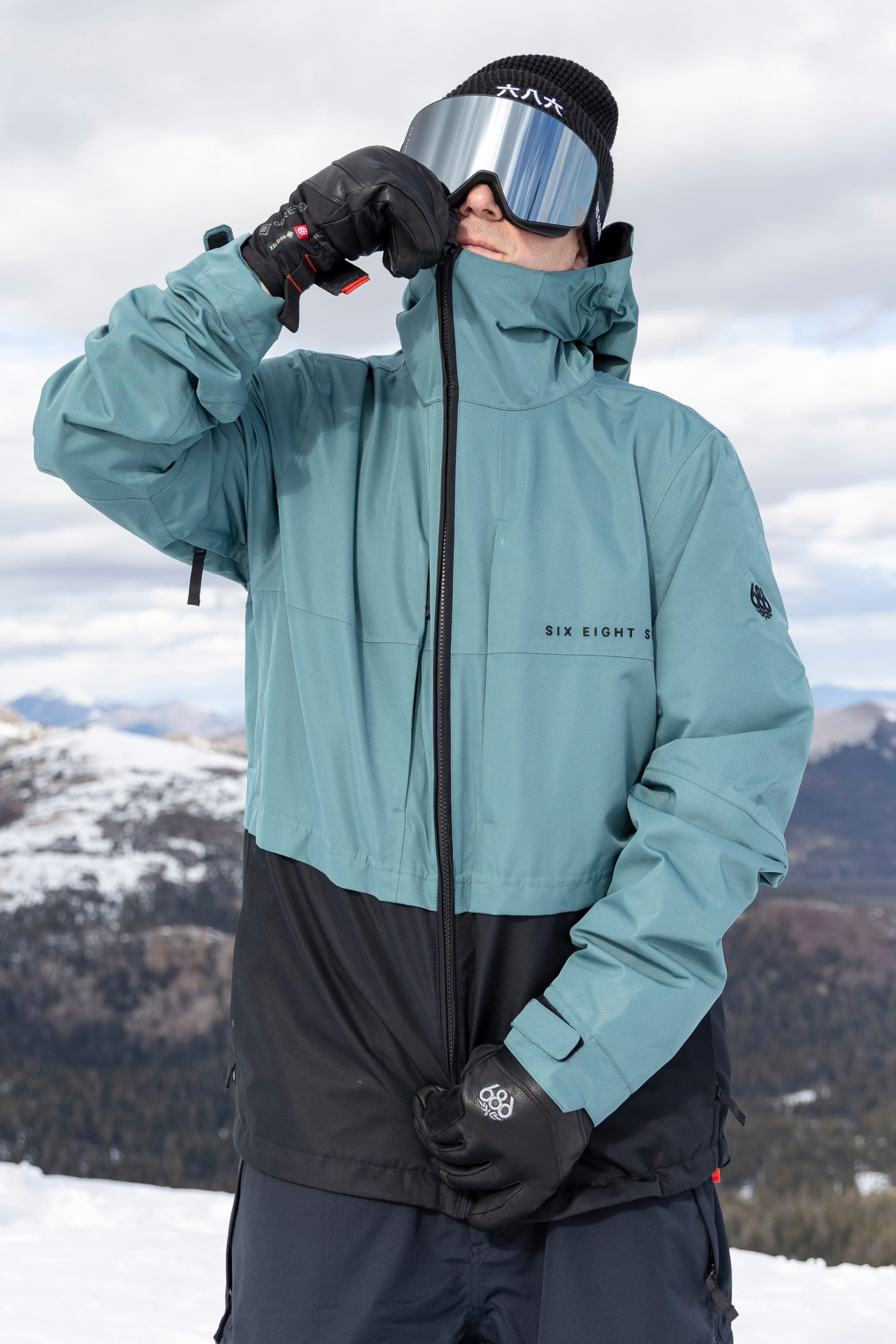 A person wearing the 686 Men’s SMARTY® 3-In-1 Form Jacket by 686, along with gloves and reflective ski goggles, is zipping up their jacket outdoors in a snowy, mountainous landscape.