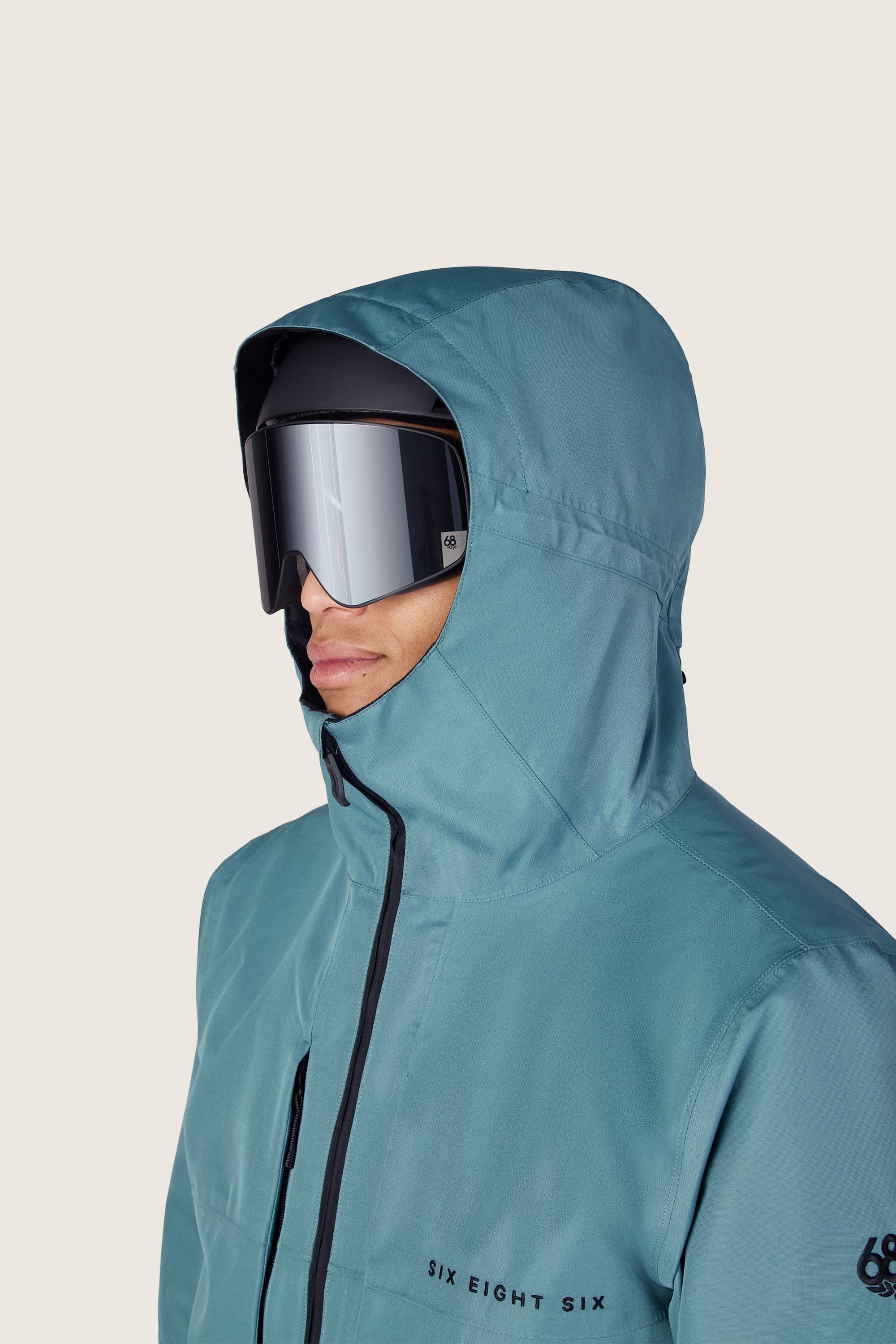 A person wearing the 686 Men’s SMARTY® 3-In-1 Form Jacket in teal and reflective ski goggles looks forward. The jacket features “SIX EIGHT SIX” text on the chest and a 686 logo on the sleeve. The background is plain and light-colored.