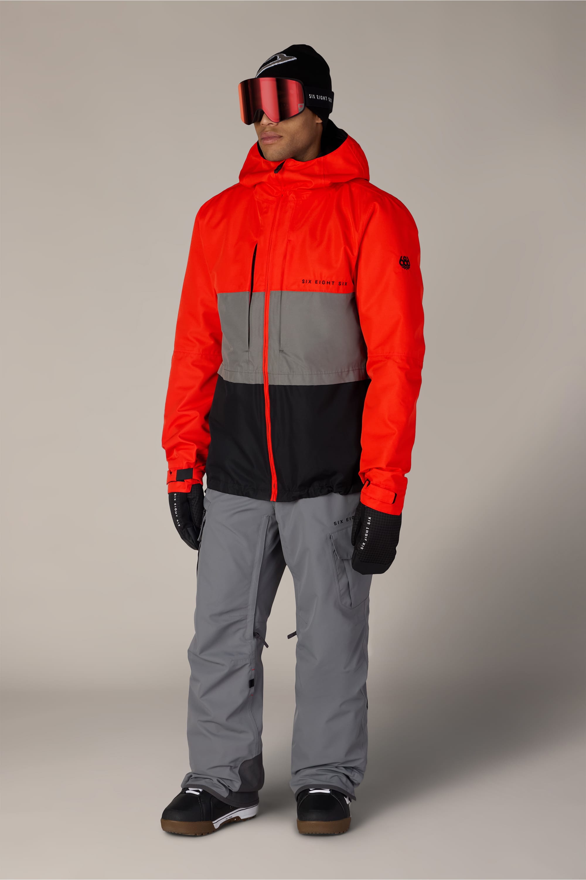 A person wearing the 686 Men’s SMARTY® 3-In-1 Form Jacket in red, gray pants, black gloves, a black hat, and red ski goggles stands against a neutral background.