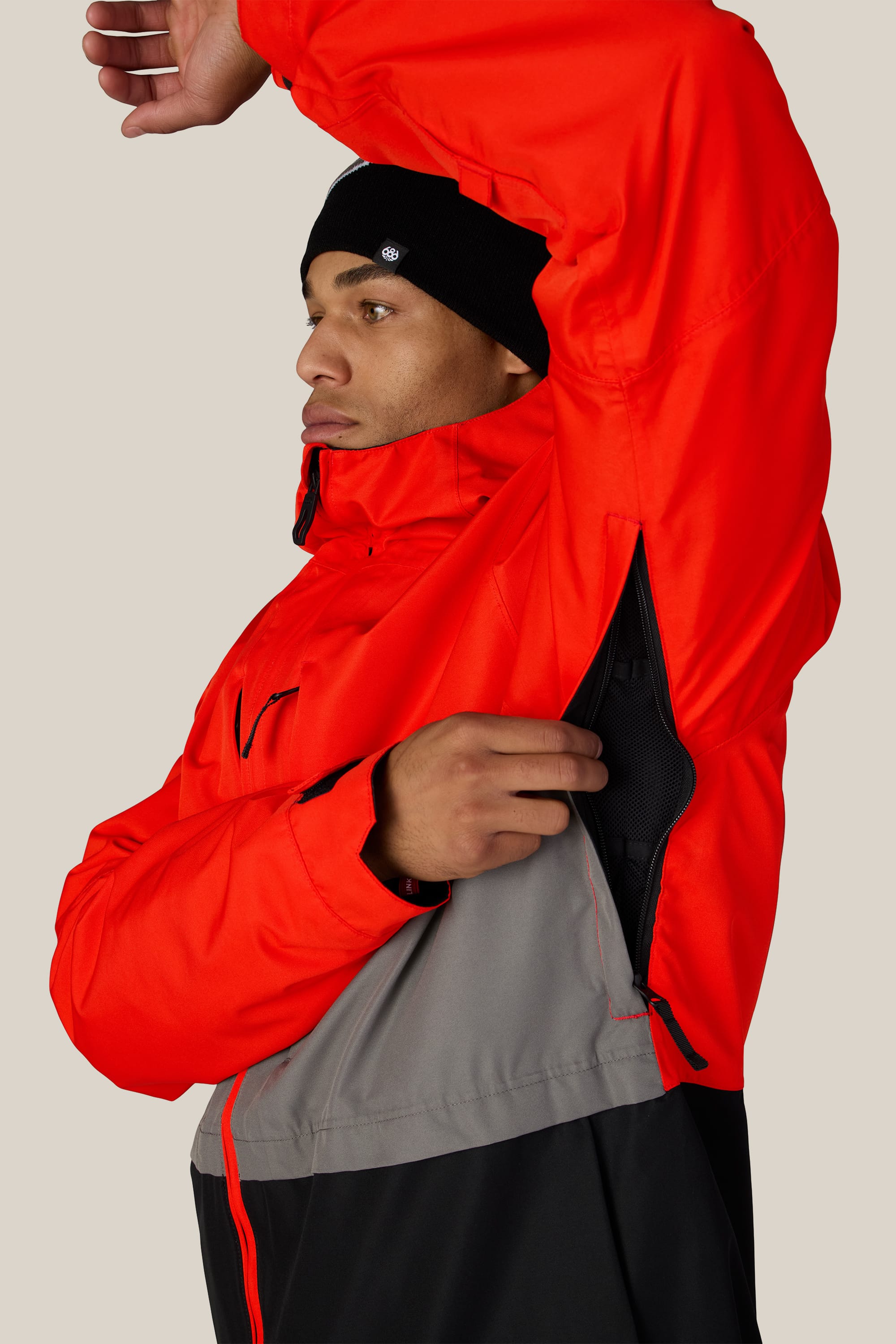 A person wearing the 686 Men’s SMARTY® 3-In-1 Form Jacket in red and gray is unzipping an underarm ventilation opening. They are also wearing a black beanie and looking to the side.