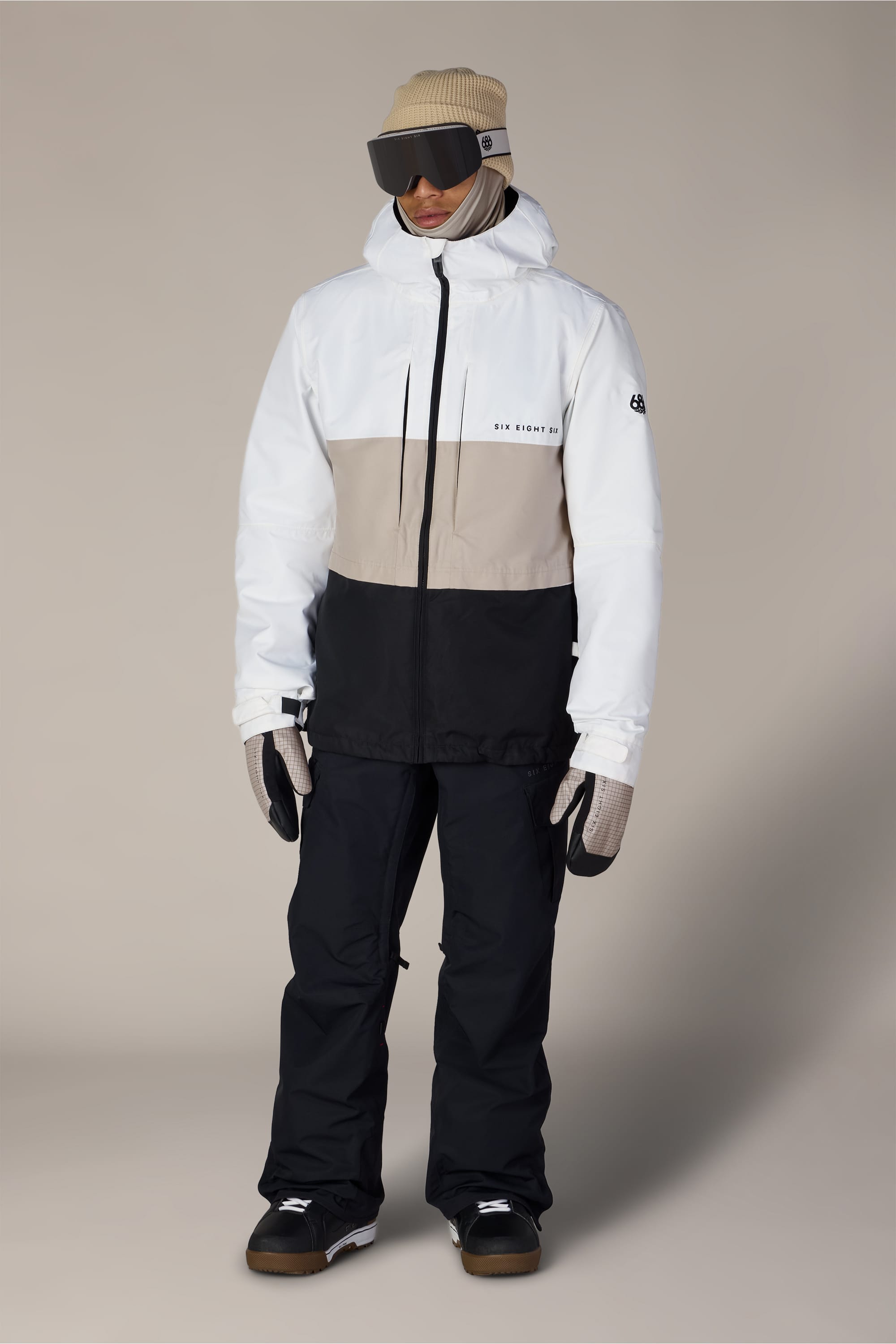A person in winter gear stands against a plain background, wearing a beige beanie, large ski goggles, black snow pants, gloves, dark boots, and the 686 Men’s SMARTY® 3-In-1 Form Jacket by 686 in white and beige.