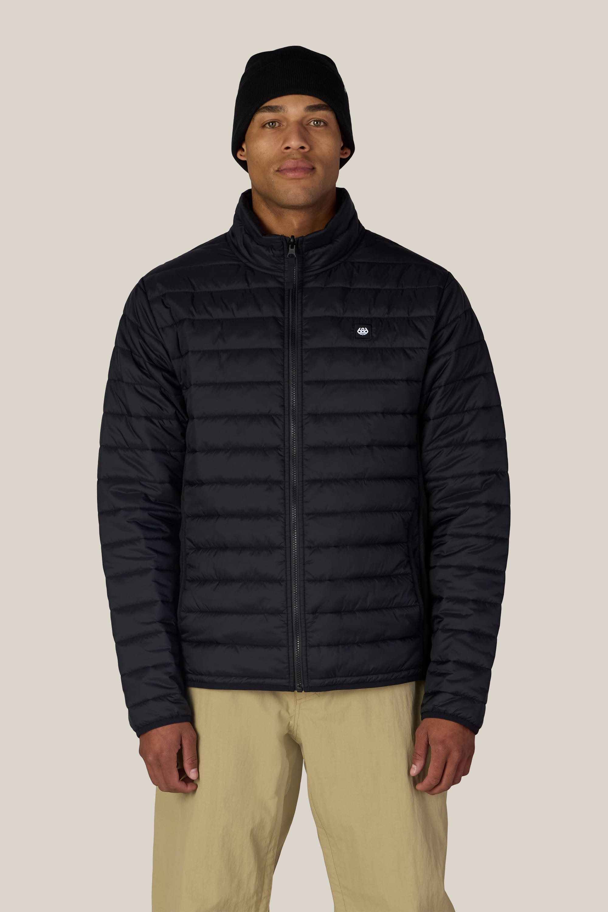 A person wearing the 686 Men’s SMARTY® 3-In-1 Form Jacket in black by 686, paired with a black beanie and beige pants, stands facing forward against a plain light background.