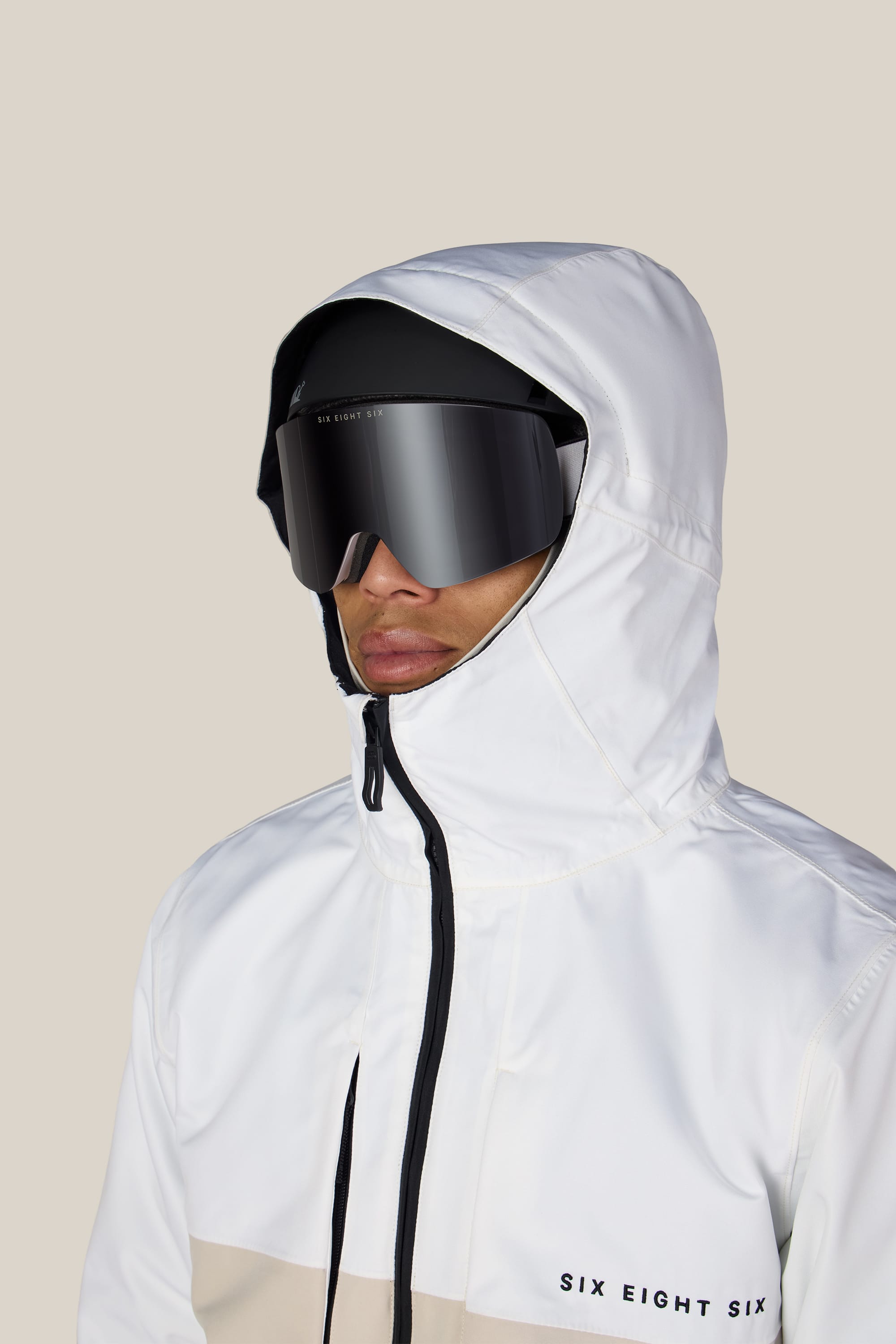 A person wearing the 686 Men’s SMARTY® 3-In-1 Form Jacket in white, featuring the 686 logo on the chest, along with large black ski goggles and a black cap, stands against a plain light background.