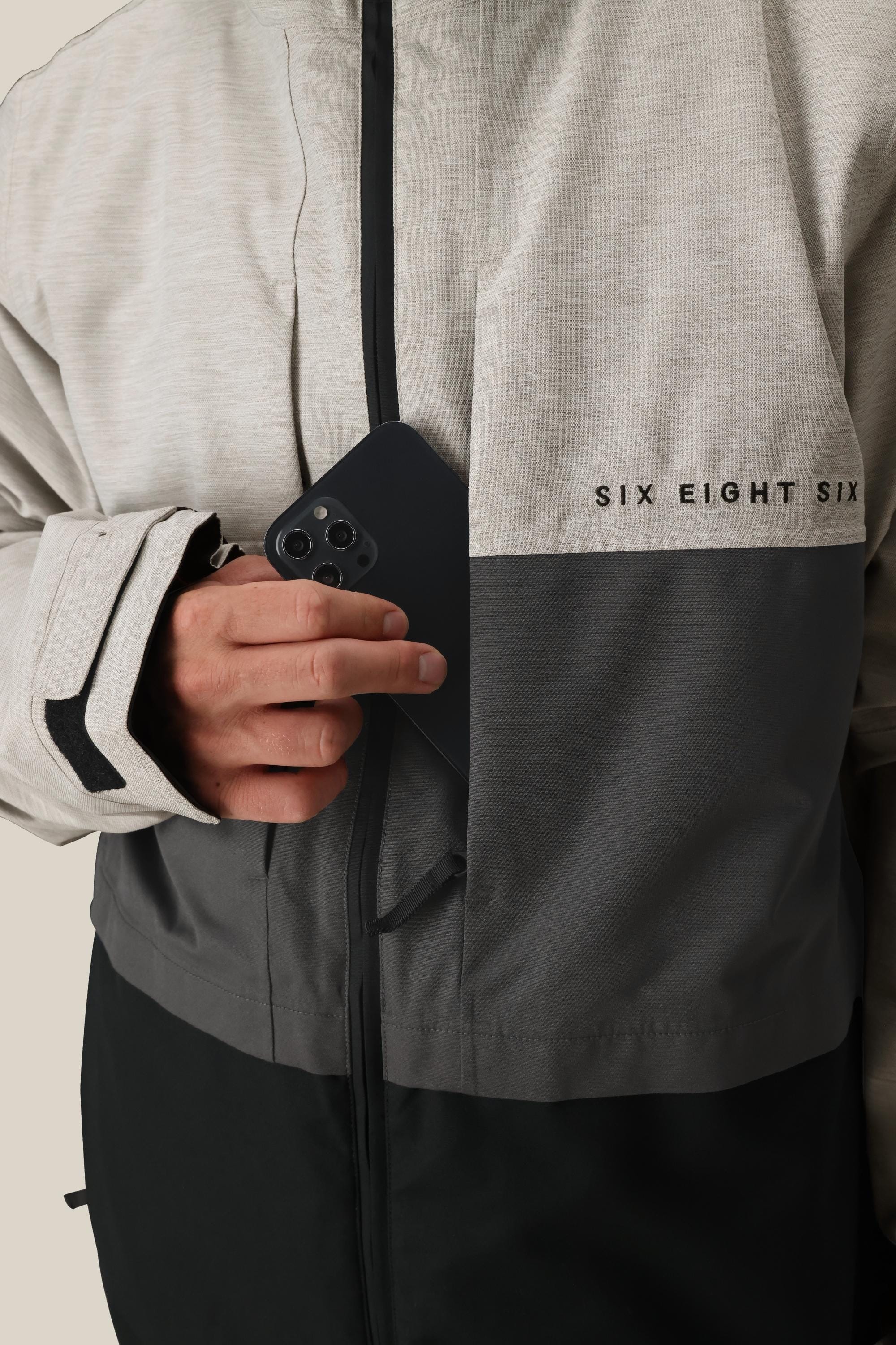 A person wearing the 686 Mens Smarty 3-In-1 Form Jacket, featuring a two-tone gray and black design with the 686 logo on the chest, slides a black smartphone into its zippered chest pocket.