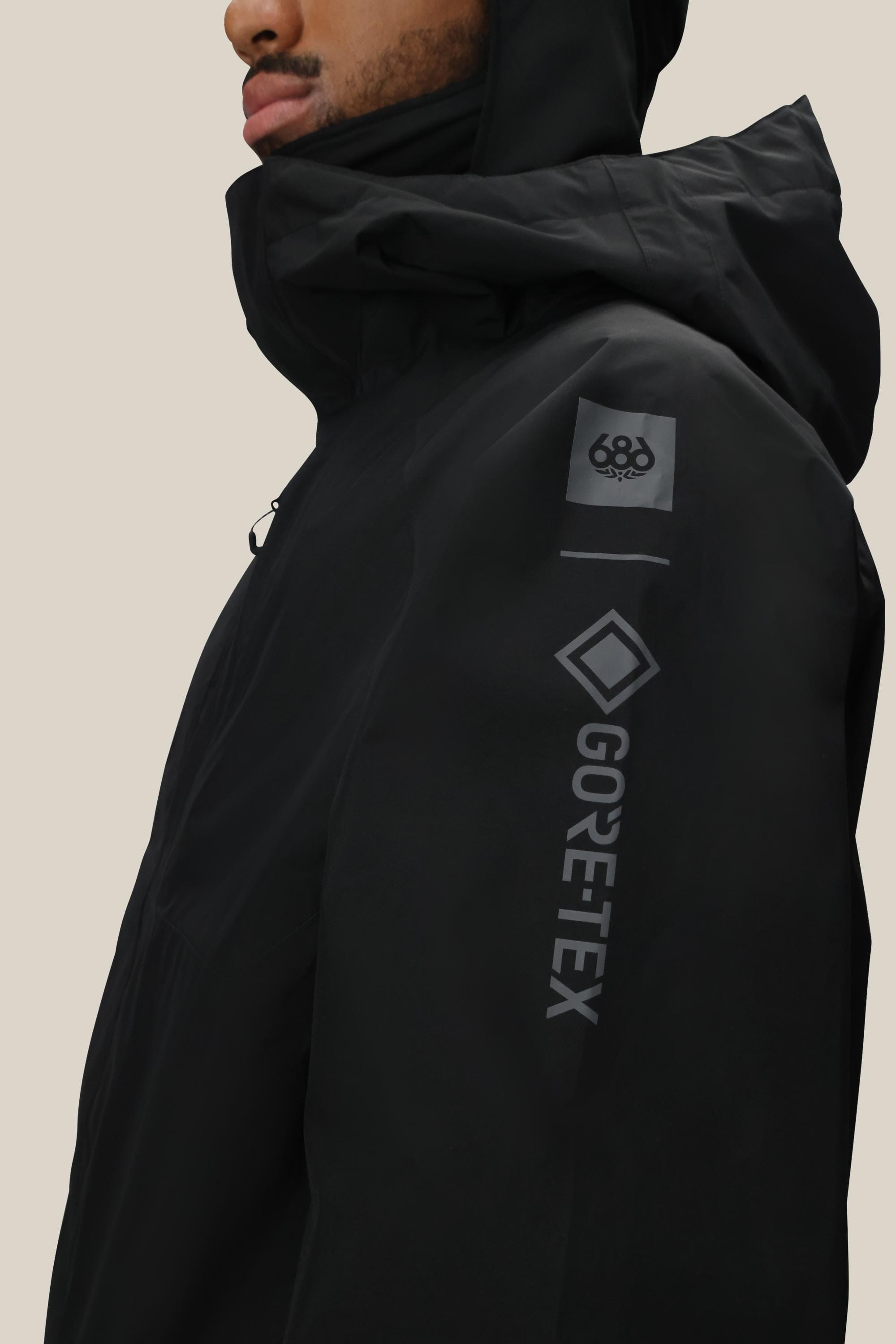 A person wearing the 686 Men’s GORE-TEX® Core Shell Jacket in black, featuring the 686 logo on the sleeve. The image displays a side view of the jacket against a plain light background.