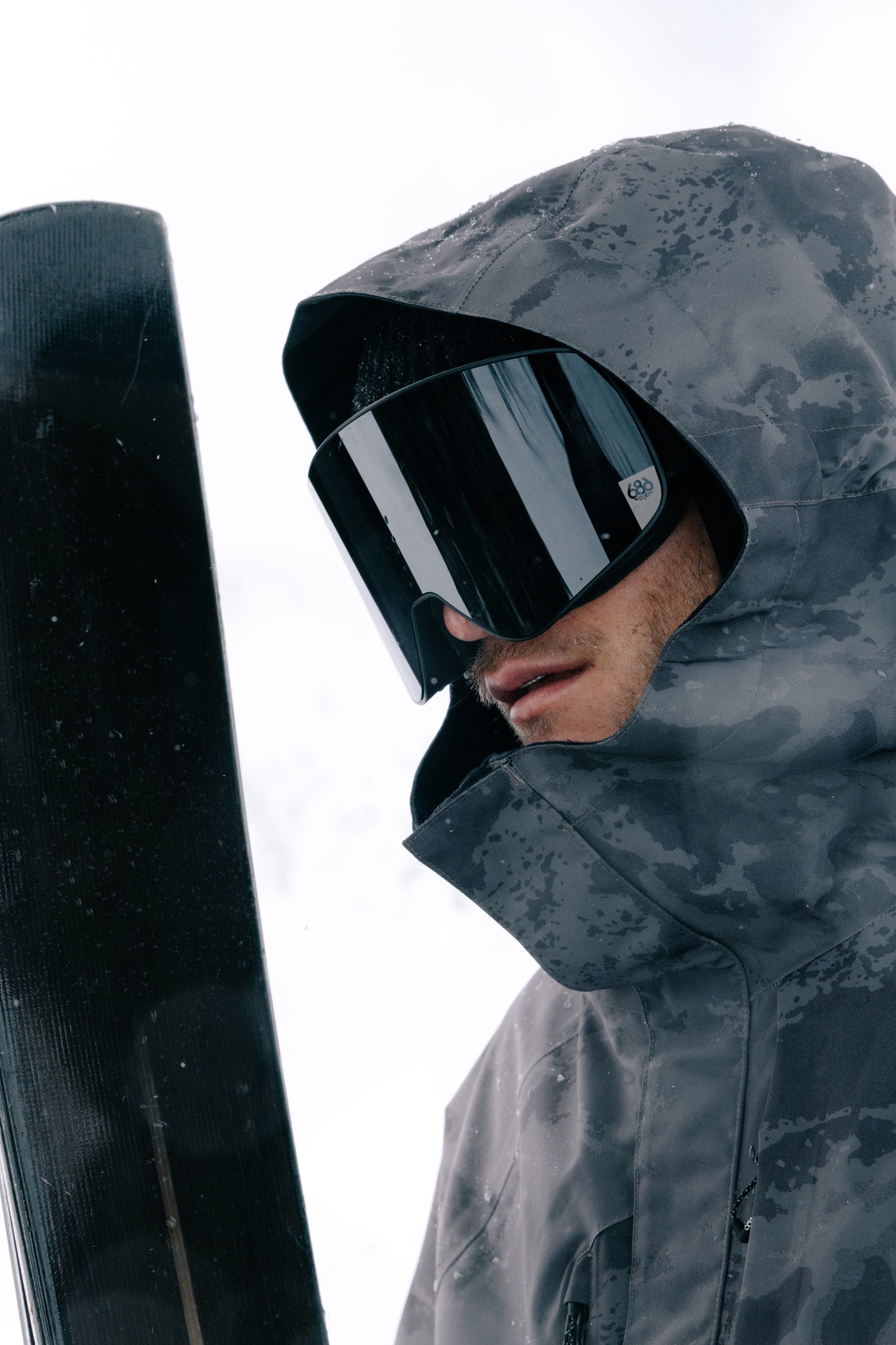 A person wearing the 686 Men’s GORE-TEX® Core Shell Jacket holds a snowboard upright in snowy outdoor conditions.