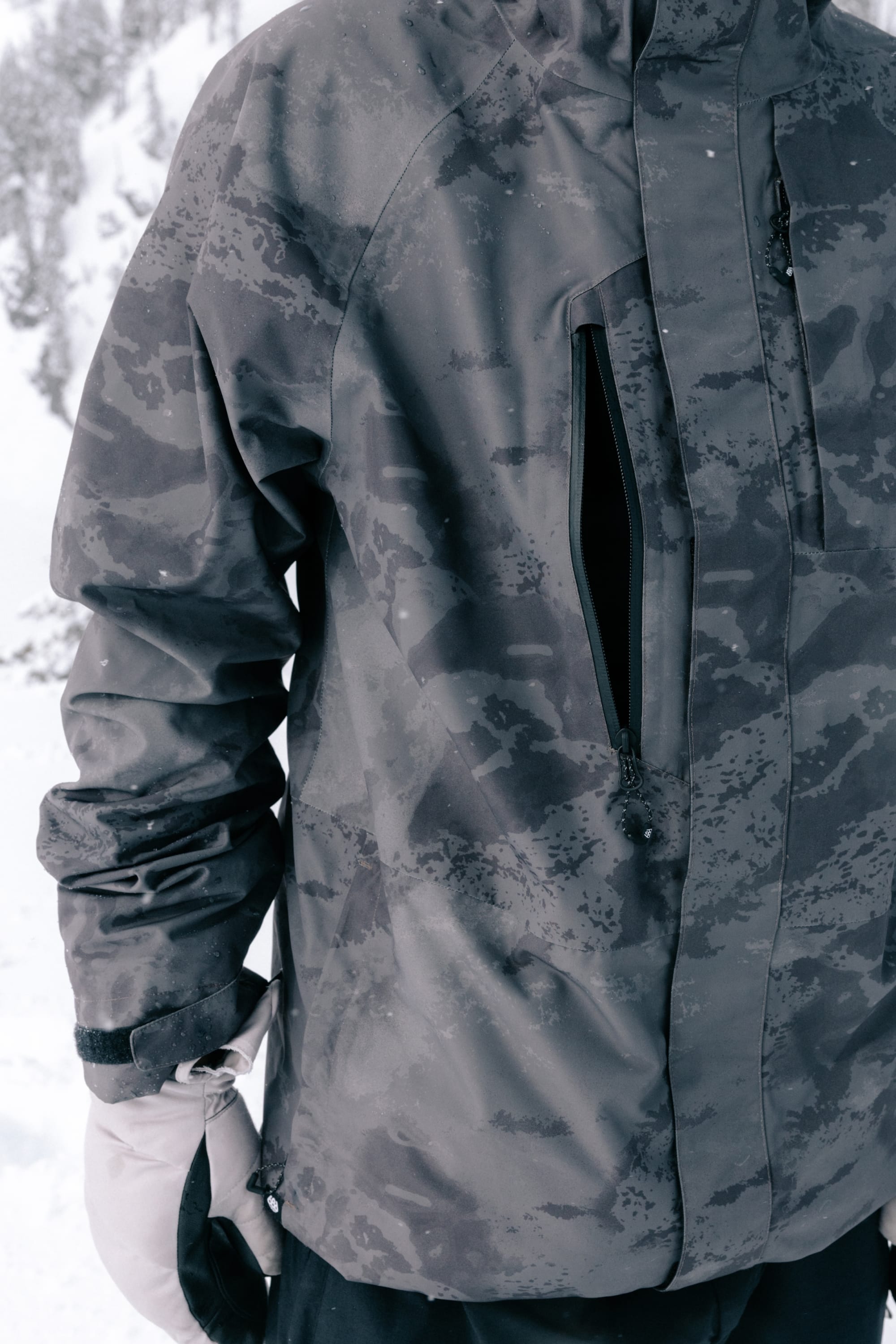 A person is wearing the 686 Men’s GORE-TEX® Core Shell Jacket in a gray camouflage pattern and white gloves, standing outdoors in a snowy landscape. The chest pocket of the 686 jacket is partially unzipped, and only the upper body is visible.