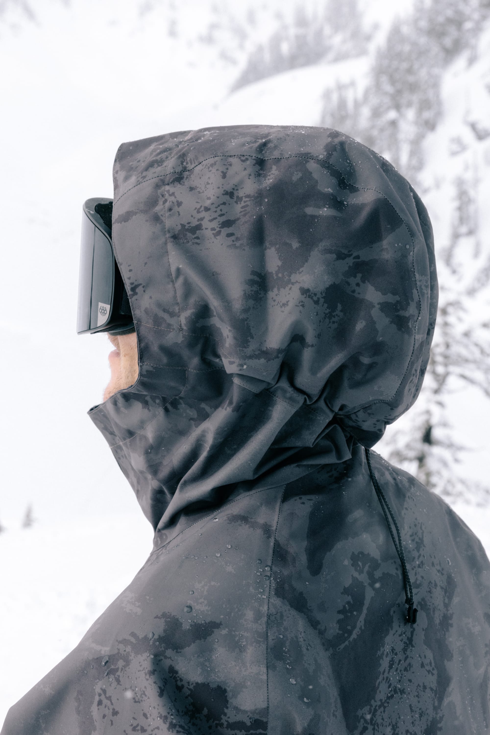 A person wearing the 686 Men’s GORE-TEX® Core Shell Jacket and ski goggles stands in a snowy outdoor setting with snow-covered trees in the background.