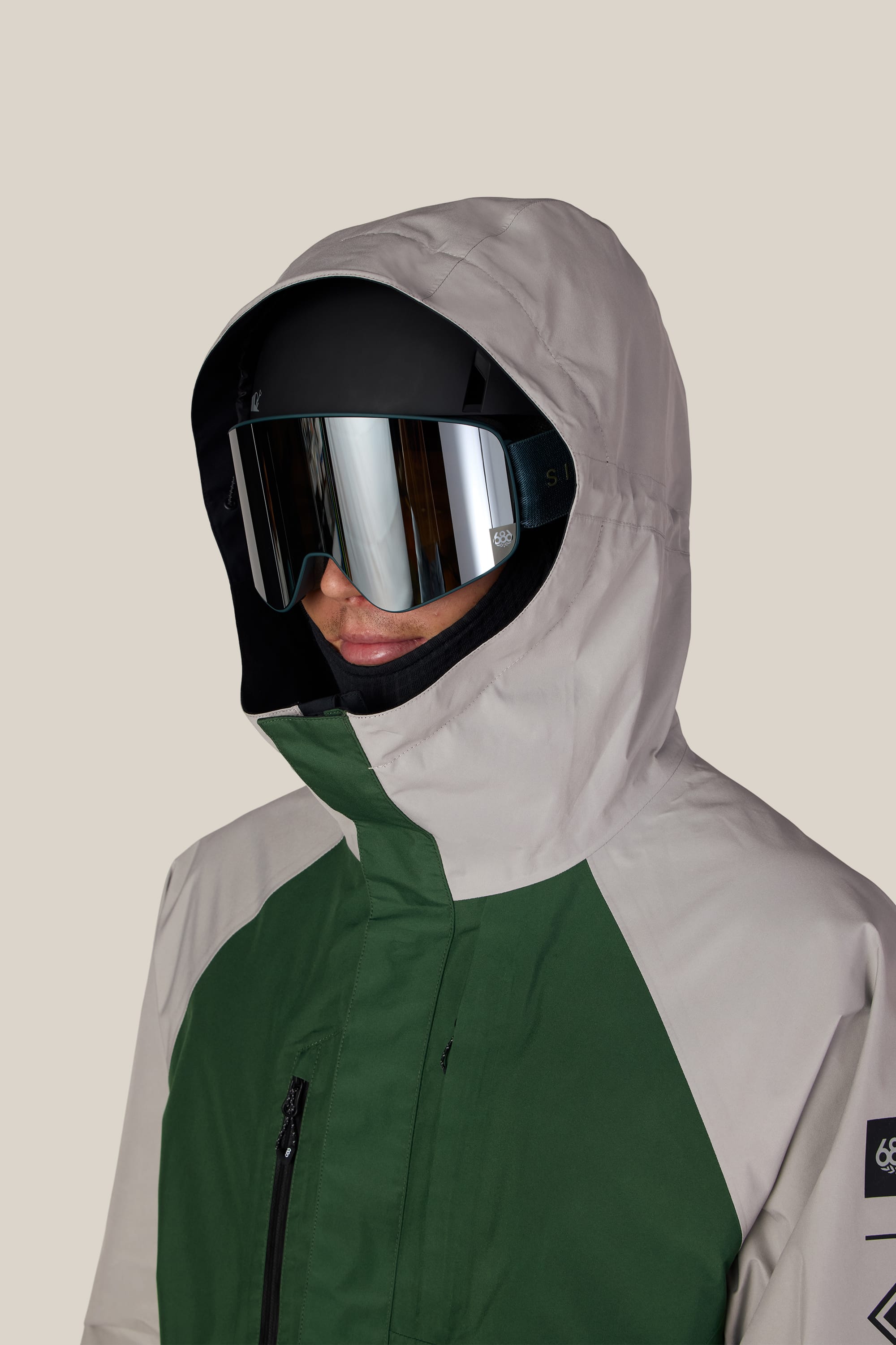 A person wearing the 686 Men’s GORE-TEX® Core Shell Jacket in green and beige, paired with reflective ski goggles, is shown from the shoulders up against a plain light background.